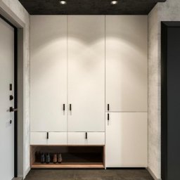 PKN Estate Sp. z o.o. - Nowoczesna, biała szafa wnękowa z drewnianą półką na buty, czarne, minimalistyczne uchwyty, szara ściana i sufit, brązowa podłoga, oświetlenie punktowe.