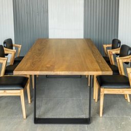 PKN Estate Sp. z o.o. - Długi, drewniany stół z czarnymi metalowymi nogami otoczony ośmioma krzesłami z jasnego drewna i czarnymi skórzanymi siedziskami na szarym tle.