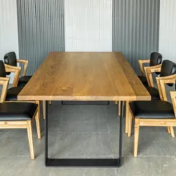 Długi, drewniany stół z czarnymi metalowymi nogami otoczony ośmioma krzesłami z jasnego drewna i czarnymi skórzanymi siedziskami na szarym tle.