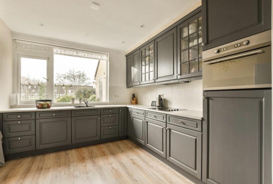 Szara kuchnia z drewnianą podłogą i dużym oknem, nad którym umieszczona jest roleta. Na blacie przy oknie stoi koszyk z owocami. Widoczne szafki z przeszkleniami.