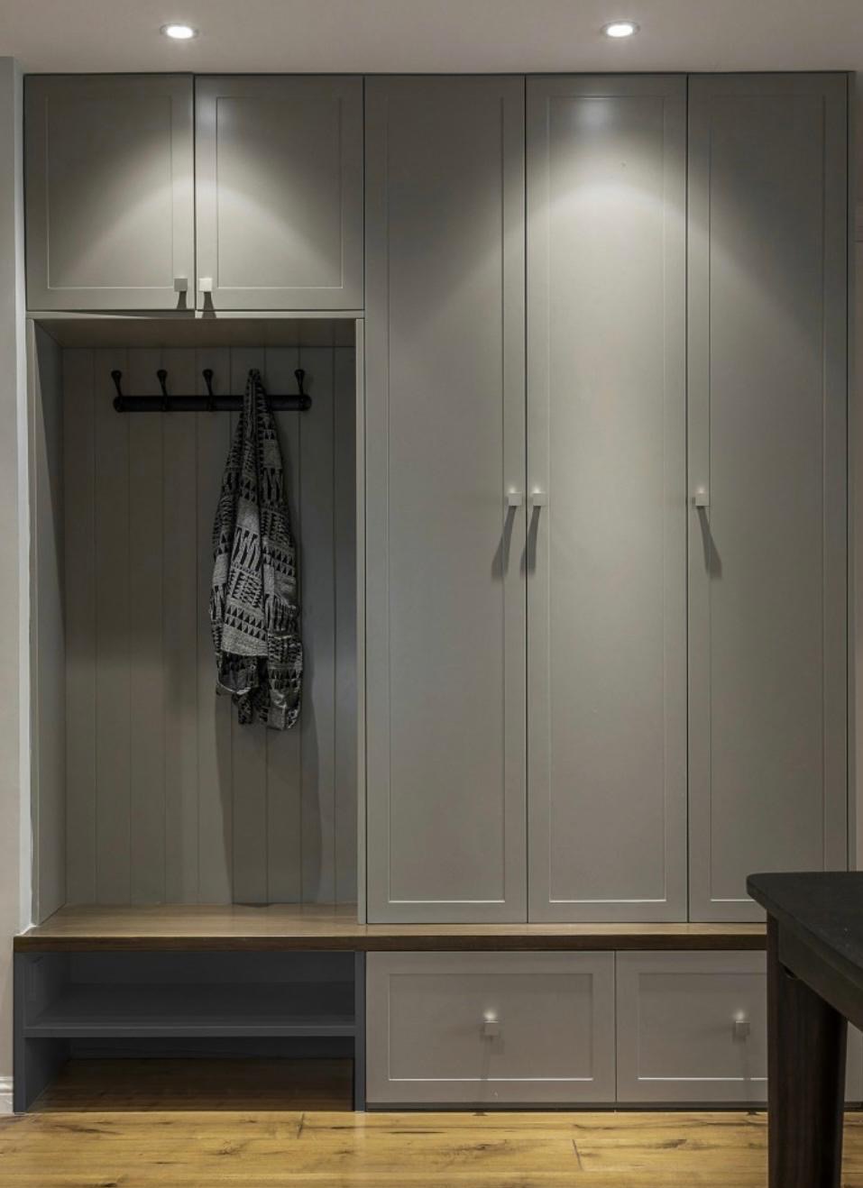 Szara szafa wnękowa z drewnianym siedziskiem i wieszakami, oświetlona punktowo, na tle drewnianej podłogi, z fragmentem ciemnego stołu po prawej stronie.