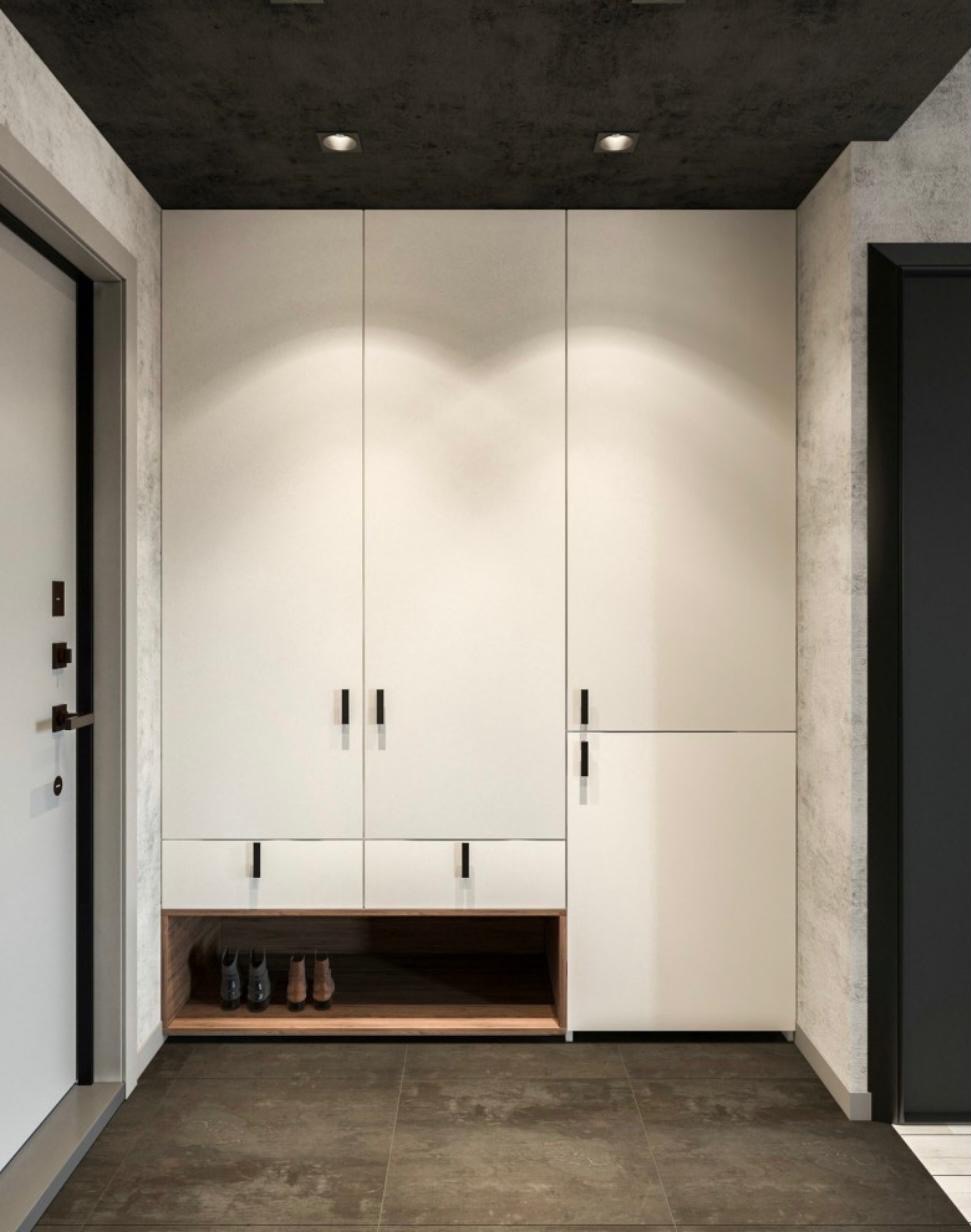 Nowoczesna, biała szafa wnękowa z drewnianą półką na buty, czarne, minimalistyczne uchwyty, szara ściana i sufit, brązowa podłoga, oświetlenie punktowe.