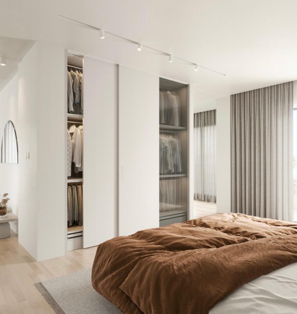 Nowoczesna szafa wnękowa z przesuwnymi drzwiami w sypialni. Jedne drzwi otwarte, ukazujące ubrania. Minimalistyczne oświetlenie szynowe. Brązowa pościel na łóżku.