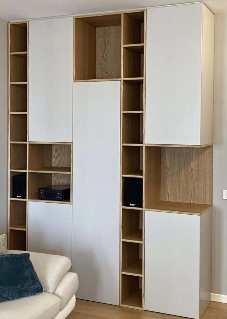 Biała szafa wnękowa z drewnianymi półkami, wkomponowana w salon, z widocznym sprzętem audio. Minimalistyczny design, jasne wnętrze, nowoczesne wykończenie.