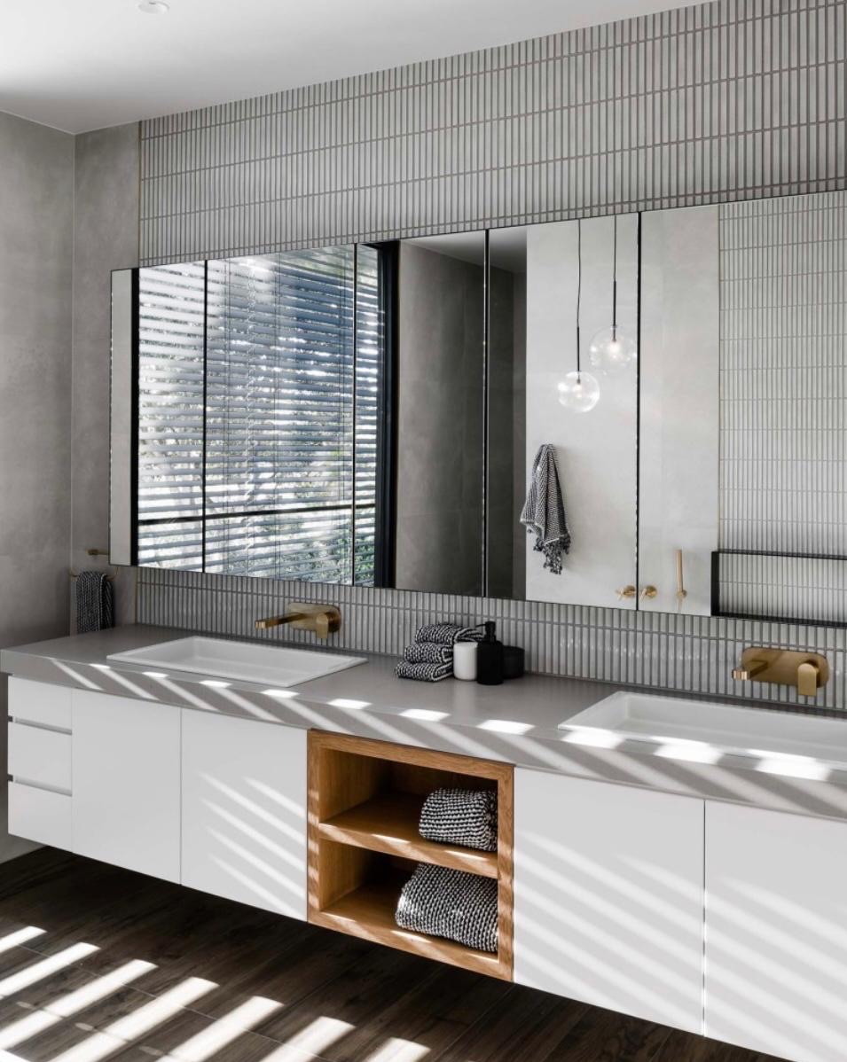 Nowoczesna łazienka z podwójną umywalką, białymi szafkami z drewnianymi półkami i lustrami na tle szarych, pionowych płytek. Minimalistyczny design z akcentami złota.