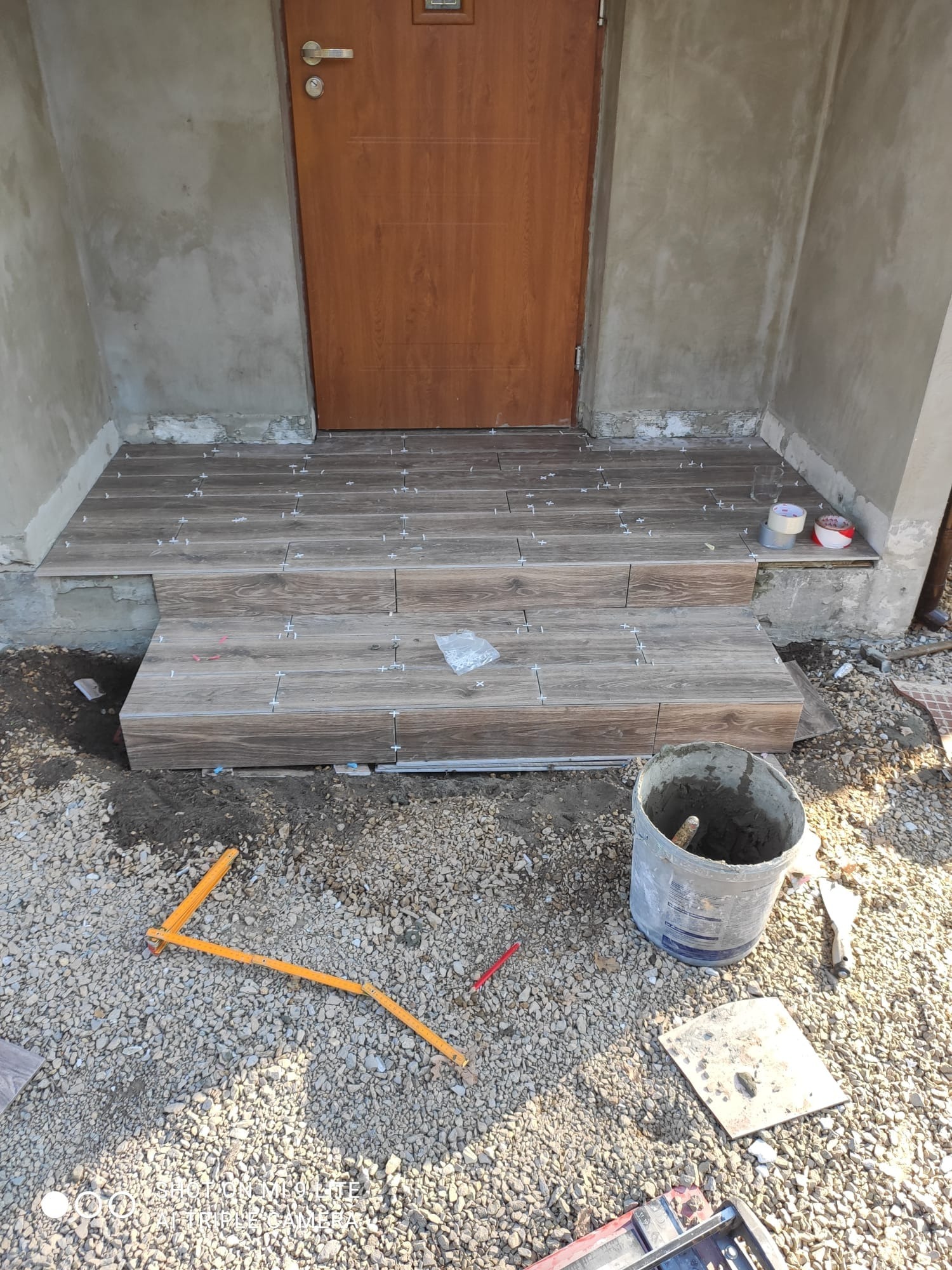 Kładzenie płytek imitujących drewno na schodach wejściowych, widoczny kubeł z zaprawą, poziomica i krzyżyki dystansowe.