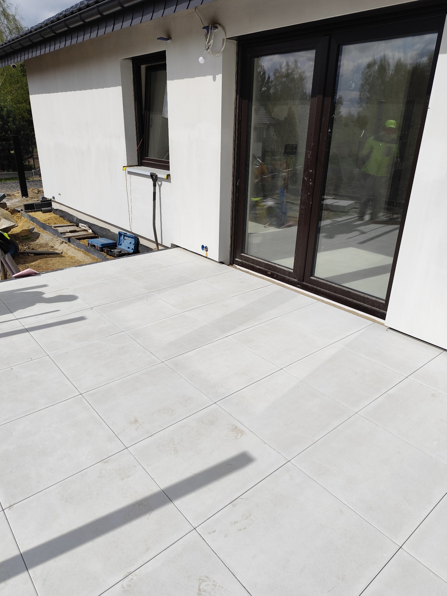 Jasnoszara kostka brukowa ułożona na tarasie przylegającym do budynku z brązowymi oknami, widoczne narzędzia i piasek w tle.