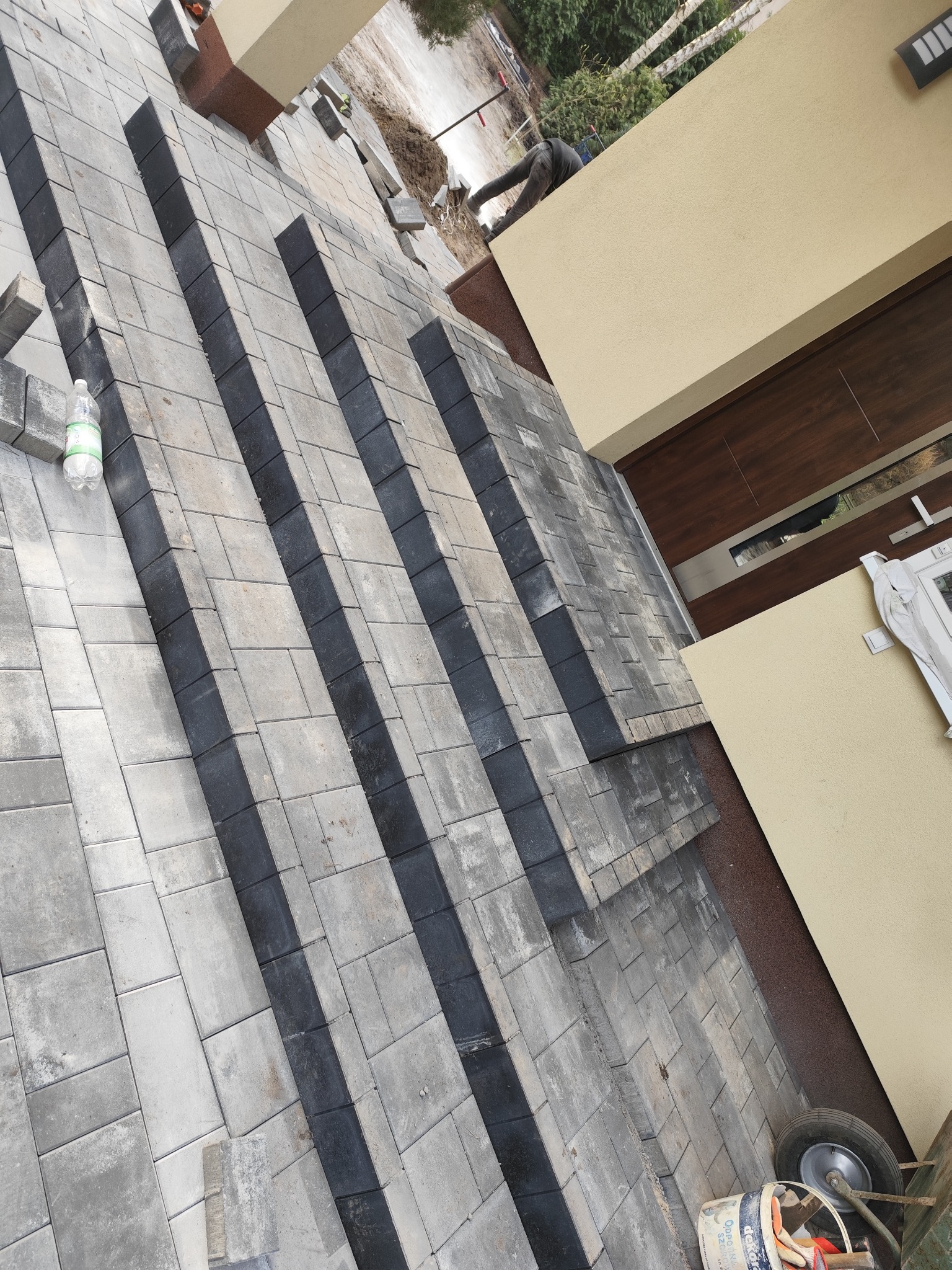 Ułożona kostka brukowa w odcieniach szarości i czerni, tworząca wzór na podjeździe, obok domu z beżową elewacją i brązowymi drzwiami. W tle widoczny pracownik oraz butelka wody i taczka z narzędziami.