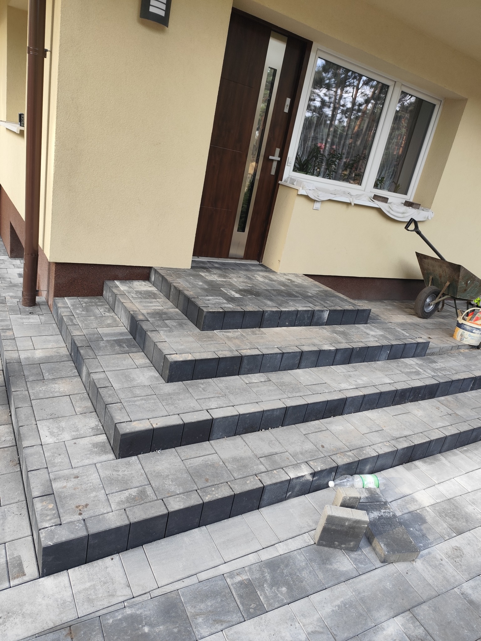 Schody wejściowe wyłożone szarą kostką brukową, z widocznymi niedociągnięciami w ułożeniu i pozostawionymi elementami roboczymi oraz butelką po napoju.