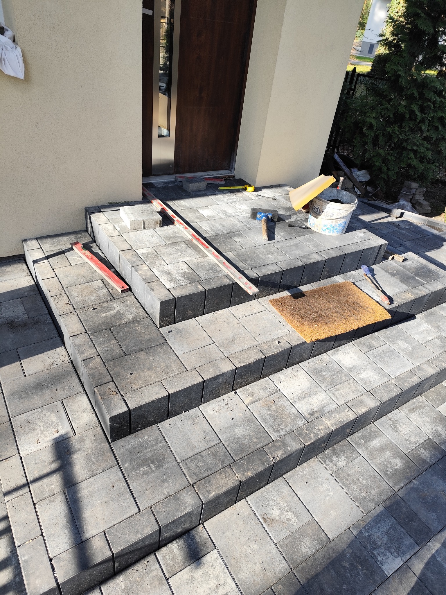 Schody wejściowe z szarej kostki brukowej w trakcie budowy, widoczne narzędzia: poziomica, młotek, wiadro z zaprawą, wycieraczka przed drzwiami.