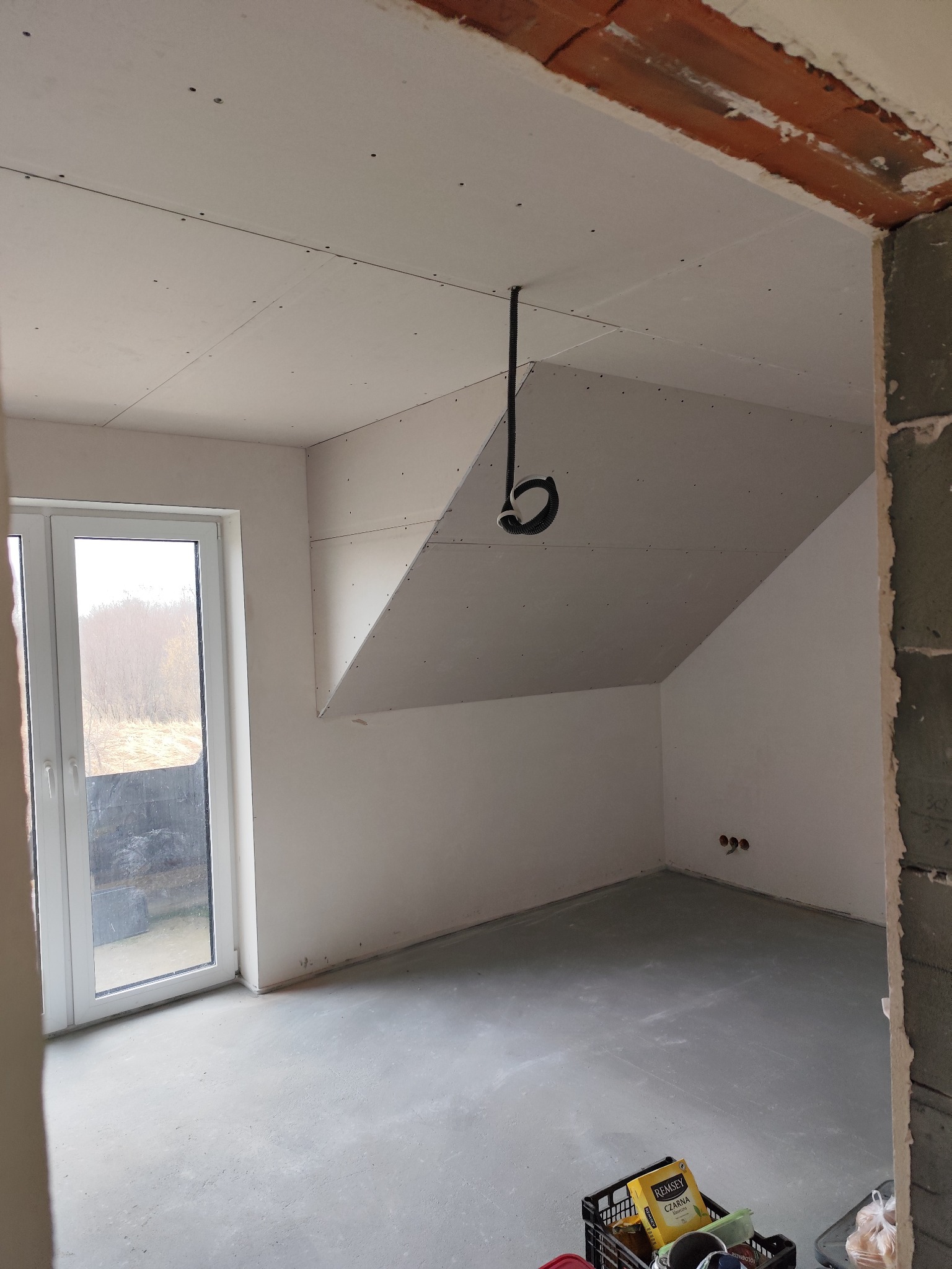 Poddasze w trakcie wykańczania: zamontowane płyty gipsowo-kartonowe, surowy beton na podłodze, widoczne przewody elektryczne i materiały budowlane w skrzynkach.