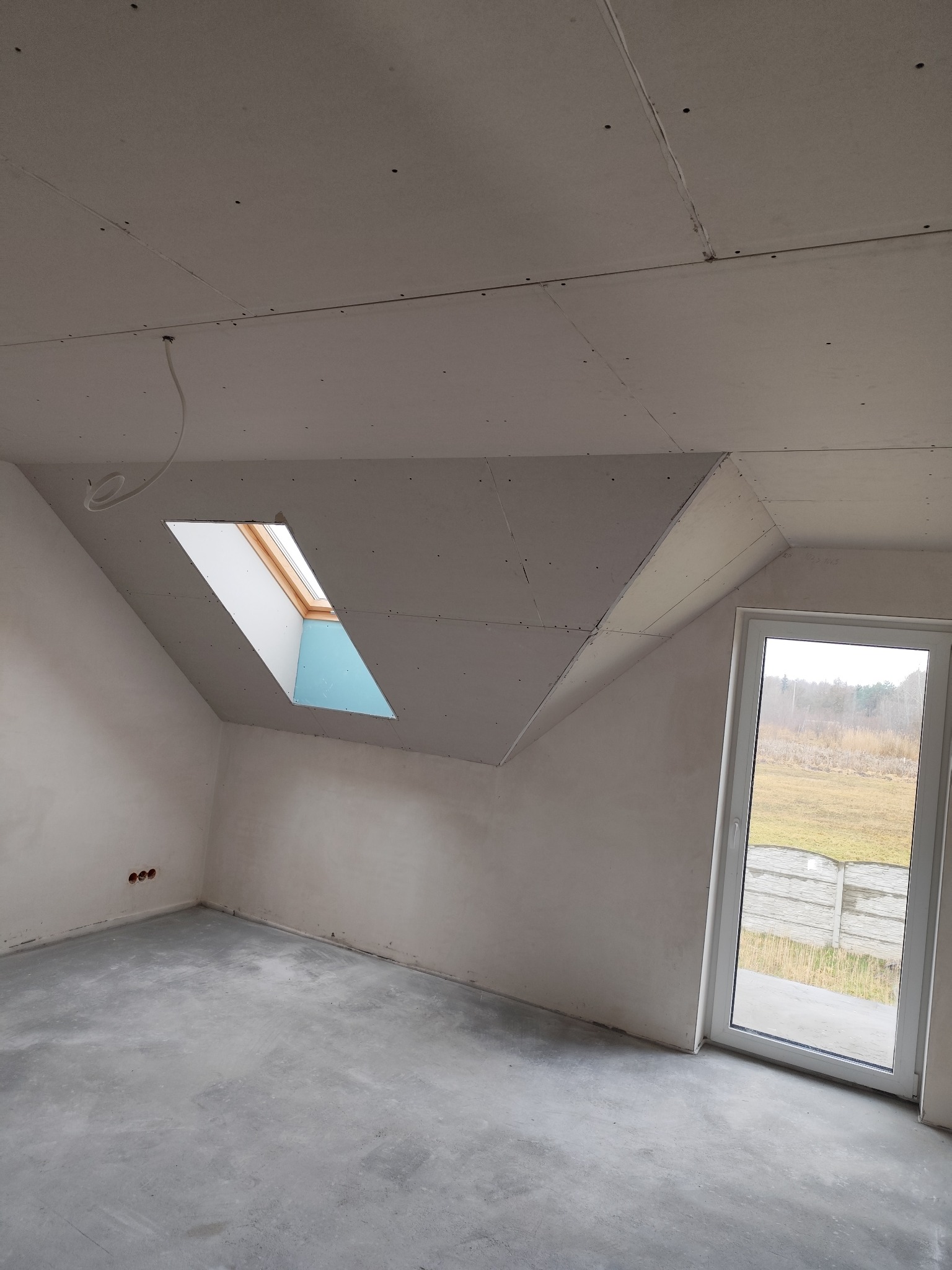 Poddasze w trakcie prac wykończeniowych: zamontowane płyty gipsowo-kartonowe na skosach dachu i suficie, widoczne okno dachowe i nieotynkowane ściany, surowa wylewka podłogowa, okno balkonowe...
