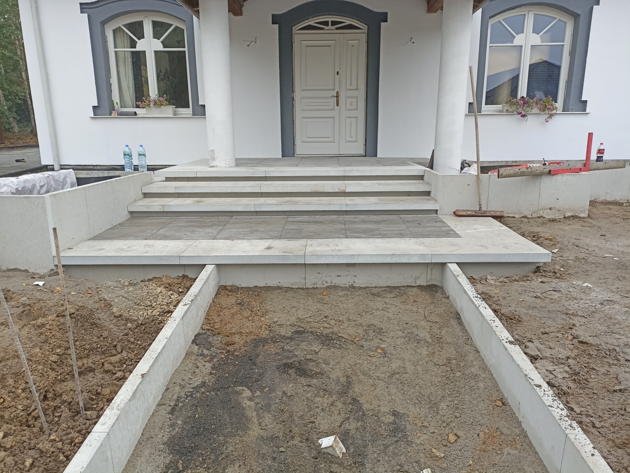 Betonowe schody wejściowe z szarymi płytami, teren wokół w trakcie przygotowania, widoczne poziomice, szczotka i butelki z wodą.