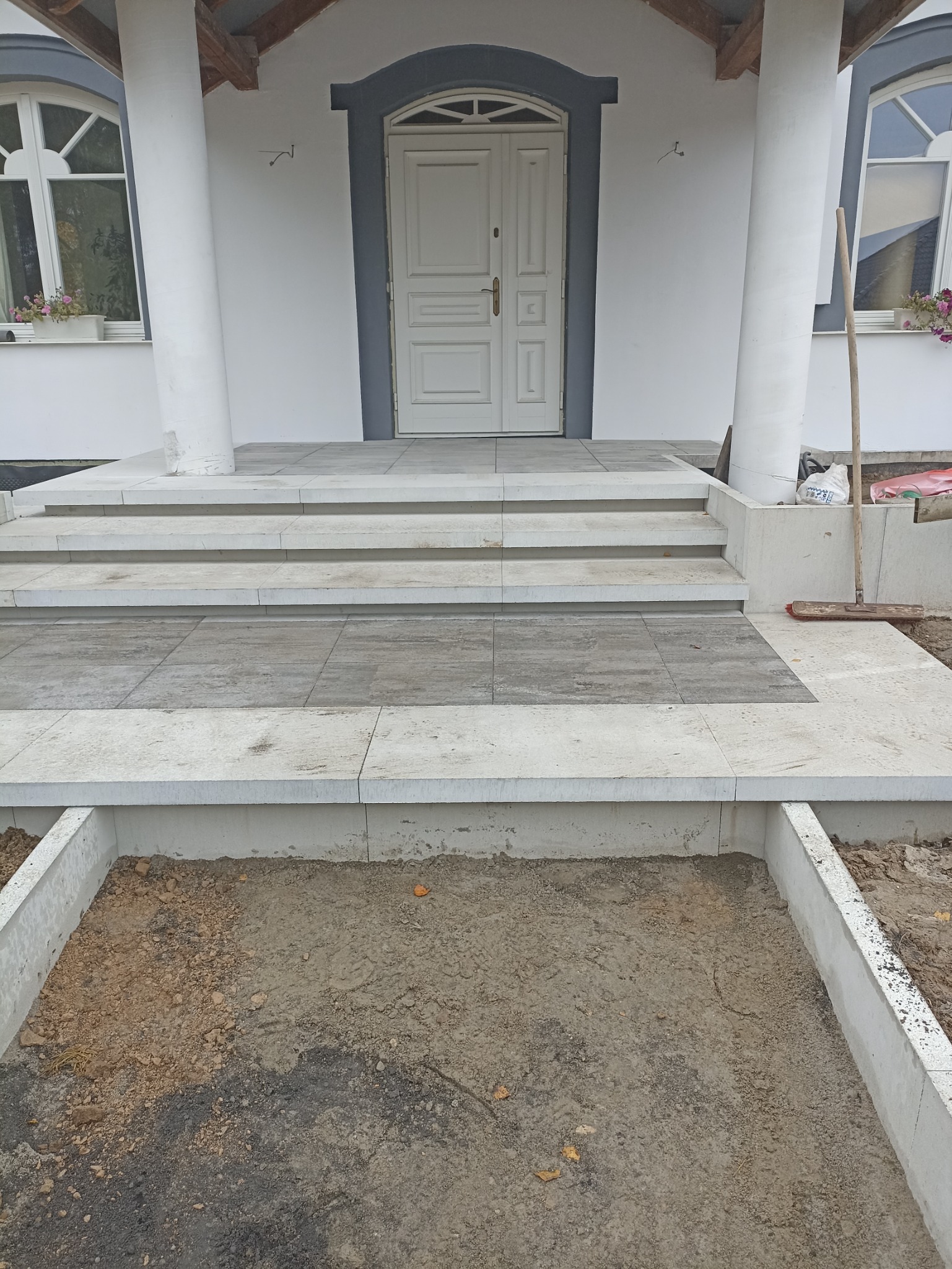 Schody wejściowe do domu z szarych płyt, częściowo ułożone, widoczny fragment przygotowanego podłoża z obrzeżami.