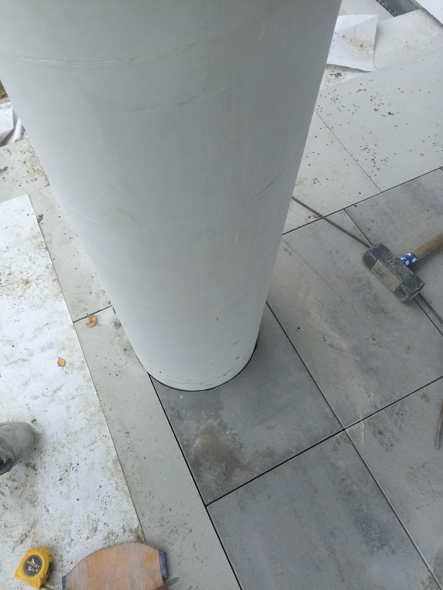 Szare płyty chodnikowe układane wokół białej, okrągłej donicy, obok leży młotek, miarka i fragment drewnianej deski.