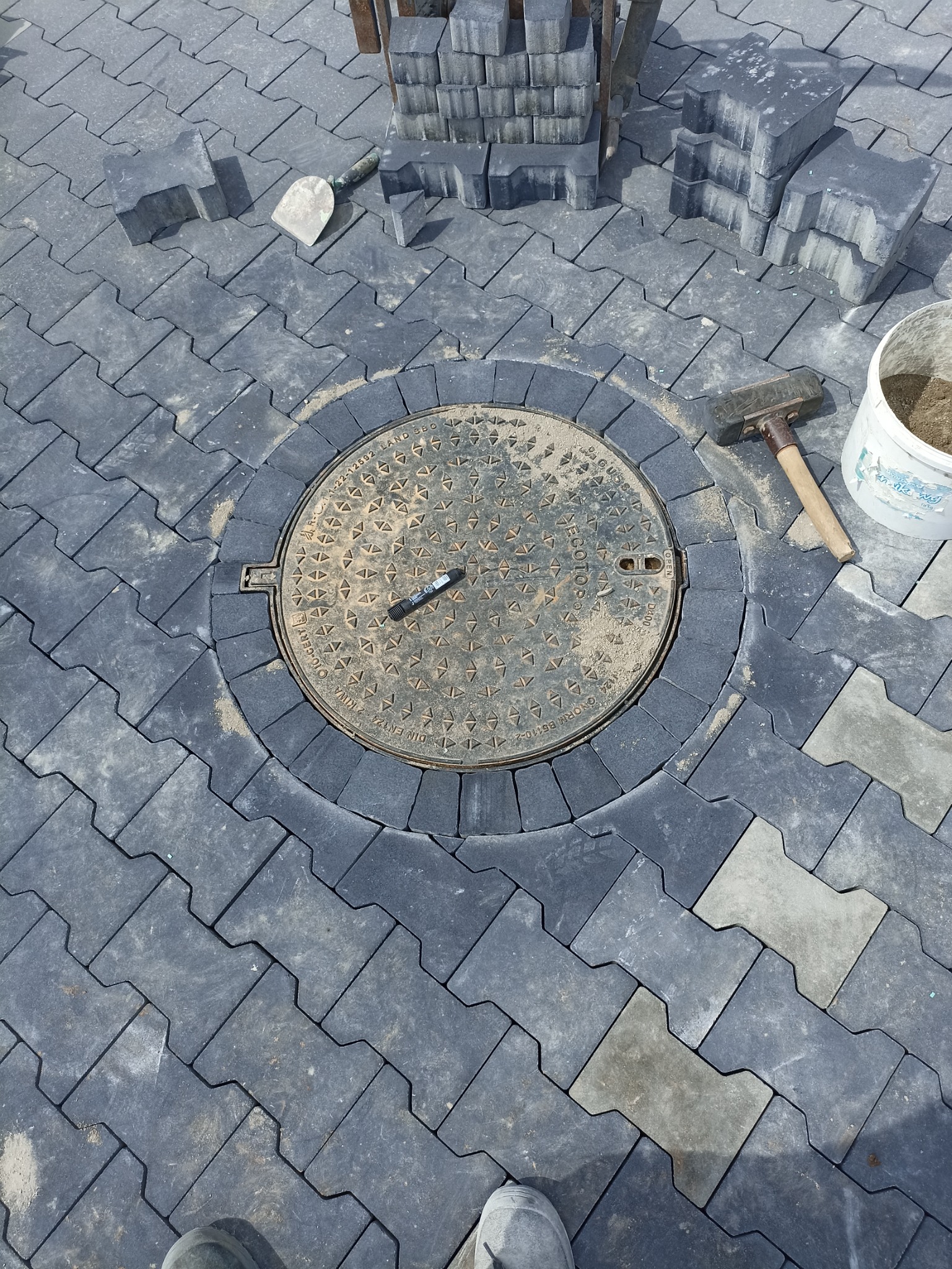 Układanie szarej kostki brukowej wokół studzienki kanalizacyjnej z narzędziami i stosem kostki w tle.