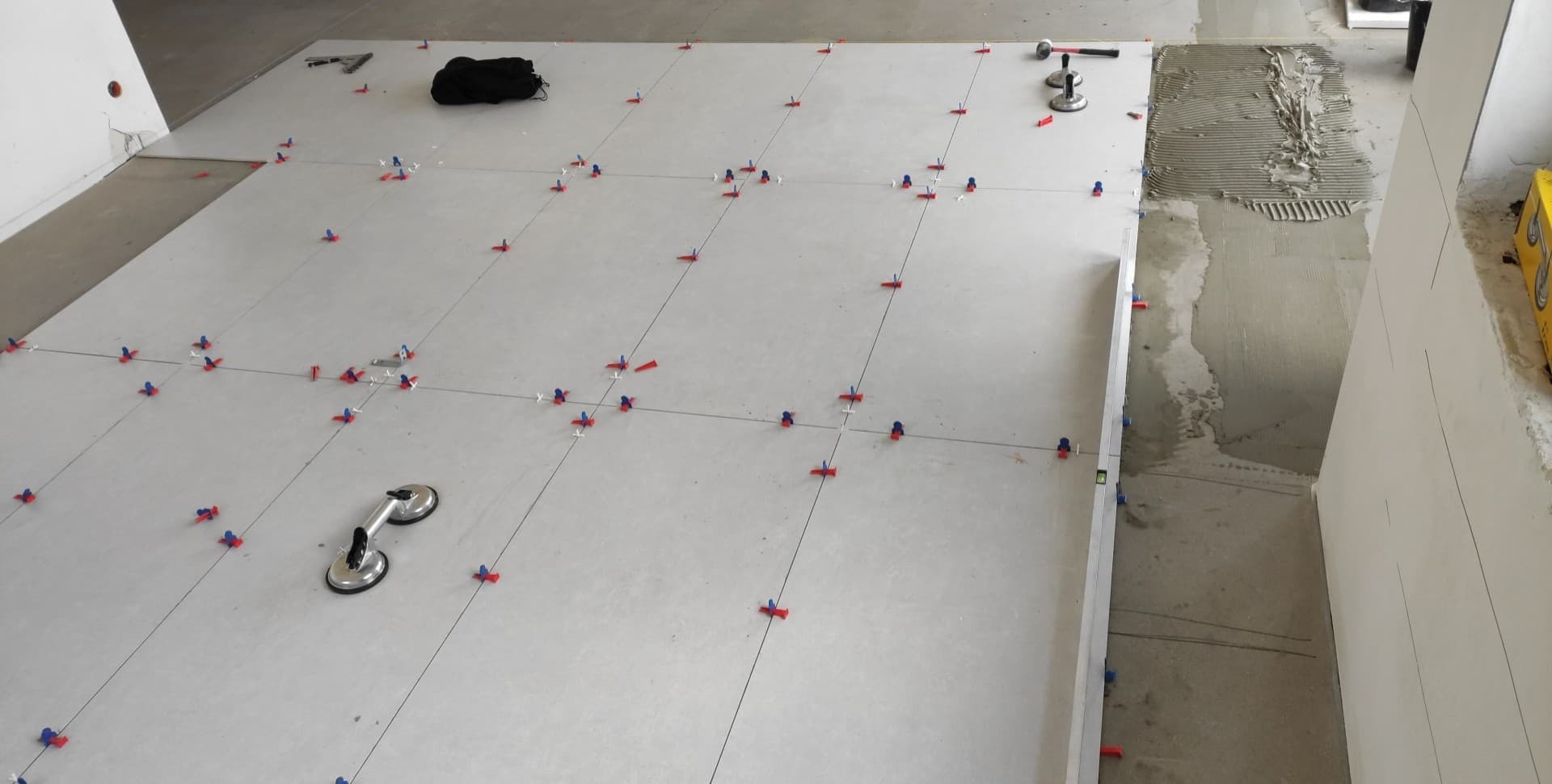 Ułożone, szare płytki podłogowe z systemem poziomowania (czerwono-niebieskie klipsy) i przyssawką do przenoszenia płytek, widoczny fragment podłoża z nałożonym klejem.