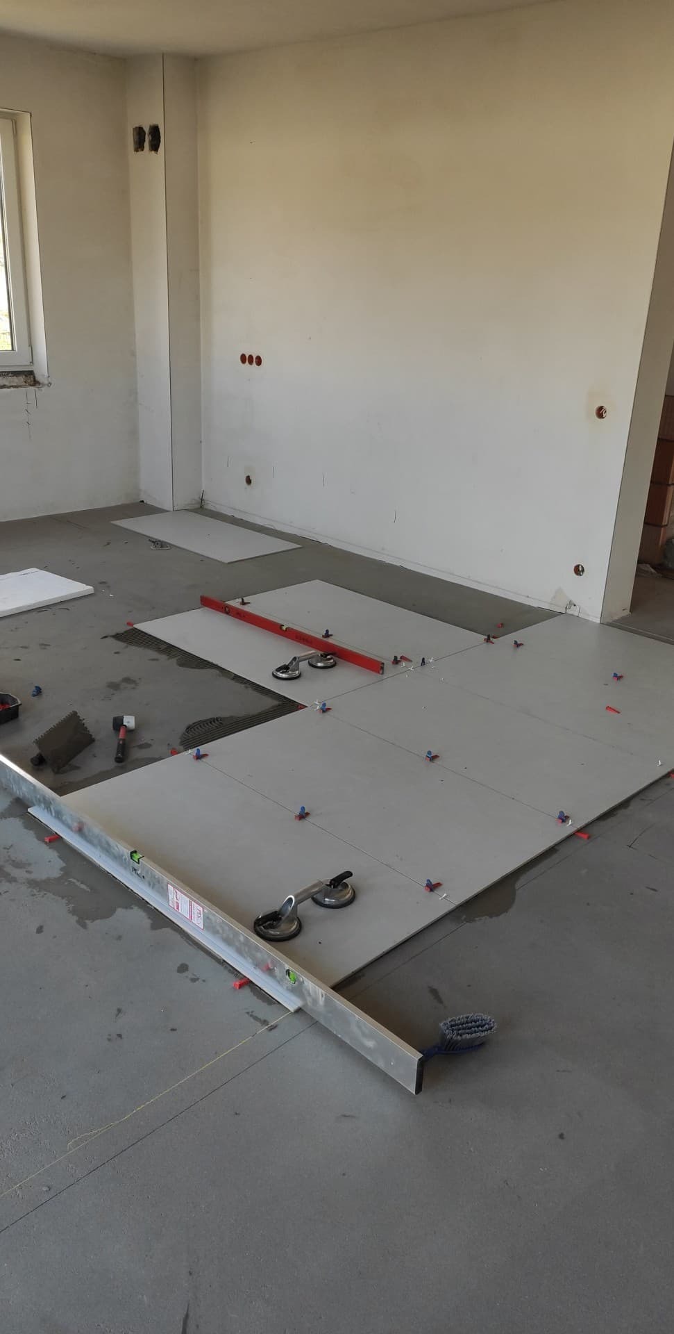 Proces układania jasnych płytek podłogowych w surowym wnętrzu, widoczne poziomice, przyssawki do płytek, krzyżyki dystansowe i ślady zaprawy.