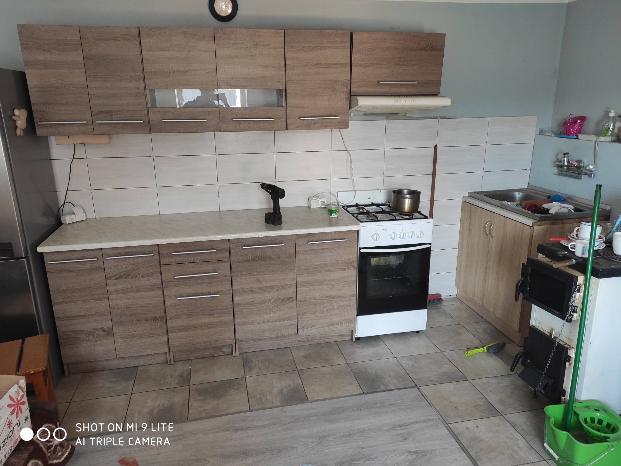 Kuchnia w trakcie remontu: widoczne brązowe szafki, białe płytki na ścianie, kuchenka gazowa z garnkiem, wiertarka na blacie i piec kaflowy obok zlewu.