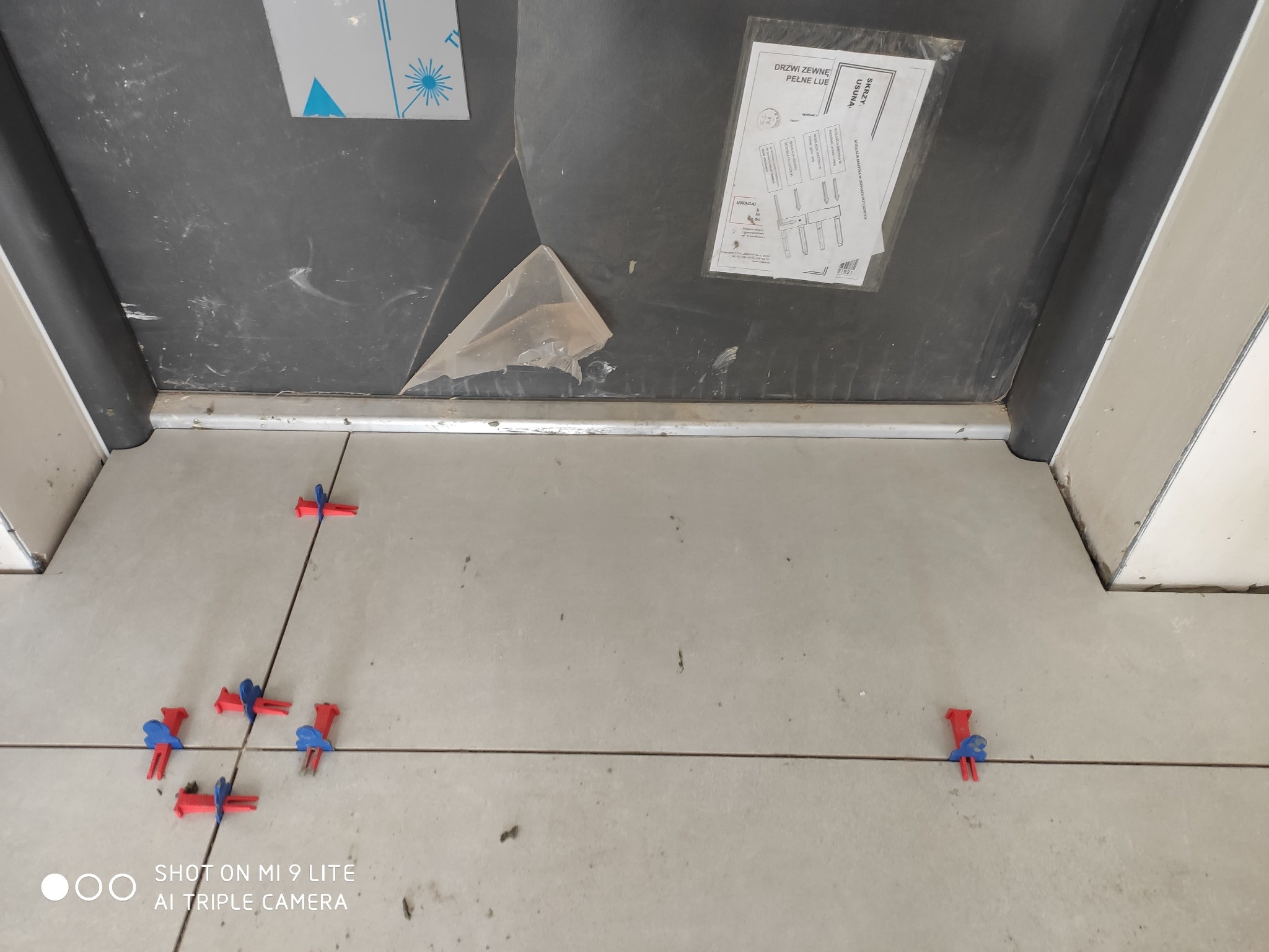Świeżo ułożone, jasne płytki podłogowe z czerwono-niebieskimi klipsami poziomującymi w spoinach, widoczne także fragmenty futryny i instrukcja montażu drzwi.