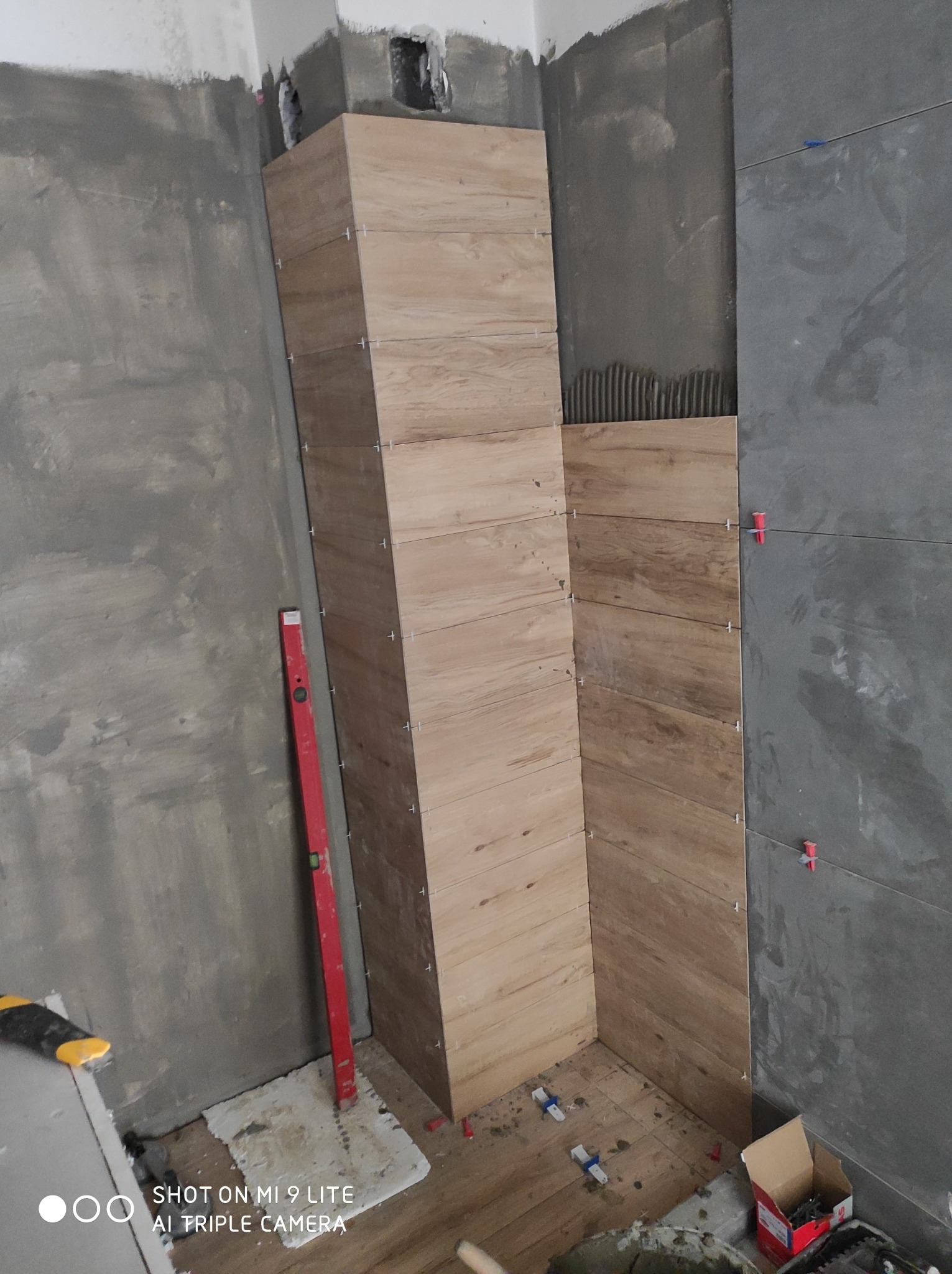 Montaż płytek imitujących drewno na ścianie w łazience z użyciem poziomicy i krzyżyków dystansowych, widoczne resztki zaprawy i narzędzia.