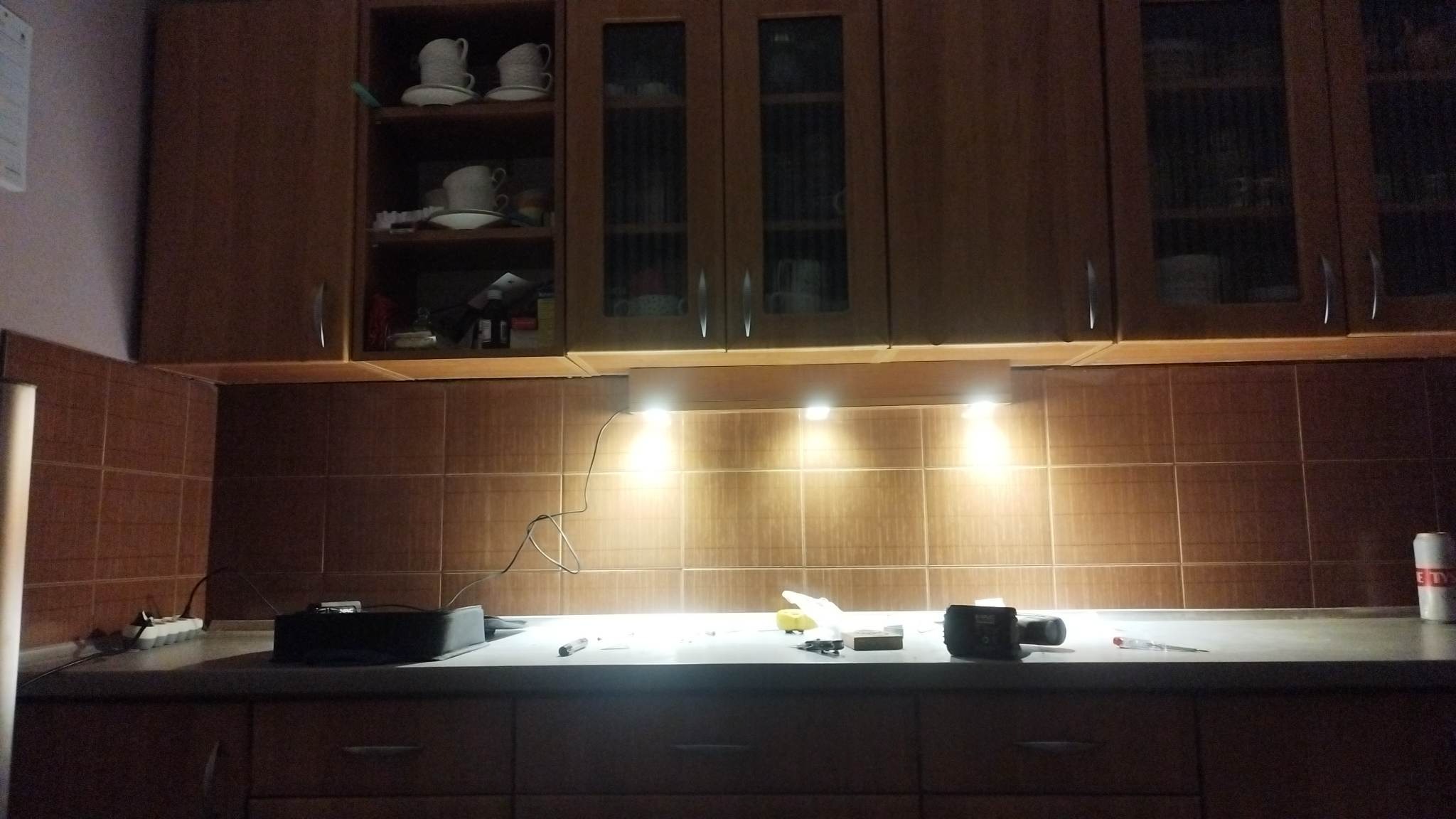 Montaż oświetlenia podszafkowego w kuchni z brązowymi płytkami, widoczne narzędzia i przewody na blacie, górne szafki z przeszklonymi frontami.