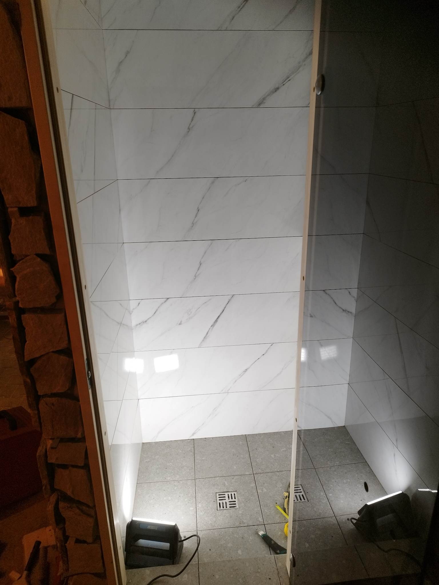 Wnętrze kabiny prysznicowej w trakcie wykańczania, widoczne białe płytki imitujące marmur na ścianach, szare płytki podłogowe z odpływem liniowym, lampa budowlana oświetlająca wnętrze i narzędzia...