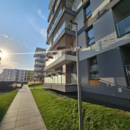 Kamera monitoringu zamontowana na słupie oświetleniowym przed nowoczesnym budynkiem mieszkalnym z balkonami i zielonym trawnikiem.