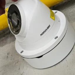 Biała kamera kopułkowa Hikvision, zdejmująca folię ochronną z obiektywu, zamontowana pod żółtą rurą na szarym, betonowym suficie.