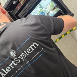 AlertSystem Krzysztof Kocot - Instalator w czarnej koszulce z logo firmy montuje system kamer. Widoczny switch, rejestrator i podgląd z kamer na ekranie w szafie rack.