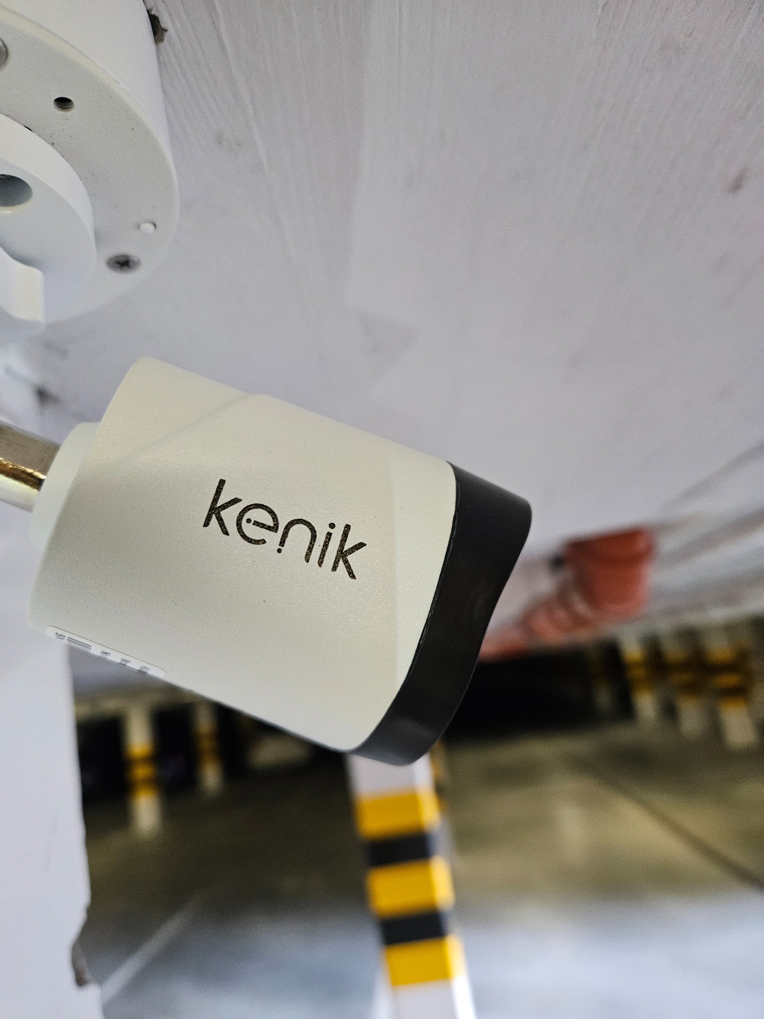 Kamera monitoringu marki Kenik zamontowana na suficie parkingu podziemnego, widoczne elementy infrastruktury parkingu w tle.