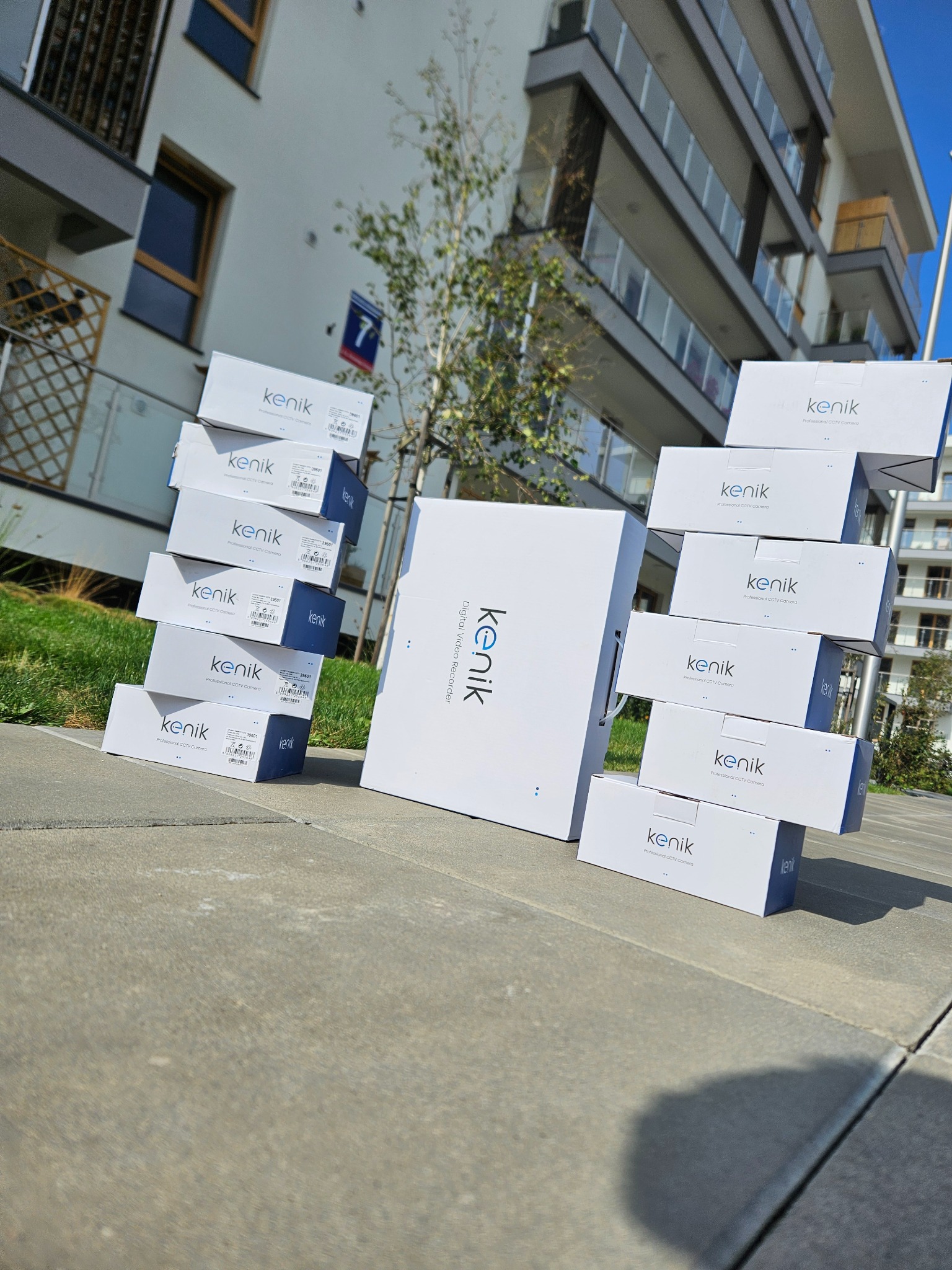Stos pudełek z kamerami CCTV i rejestratorem marki Kenik na tle nowoczesnego budynku mieszkalnego z numerem 7, sugerujący dostawę sprzętu do monitoringu.