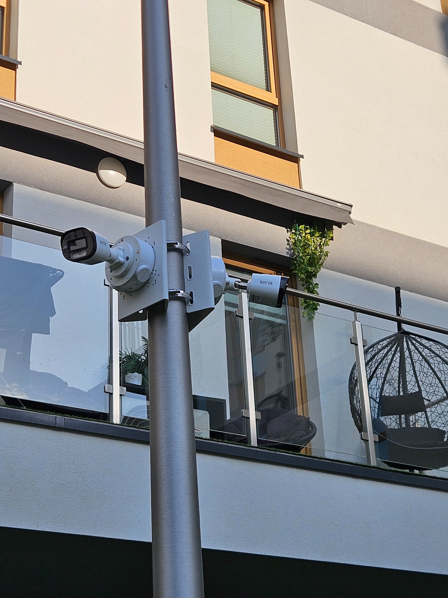 Dwie kamery przemysłowe zamontowane na metalowym słupie, skierowane w różnych kierunkach, widok z dołu na balkon z krzesłem wiszącym i roślinami.