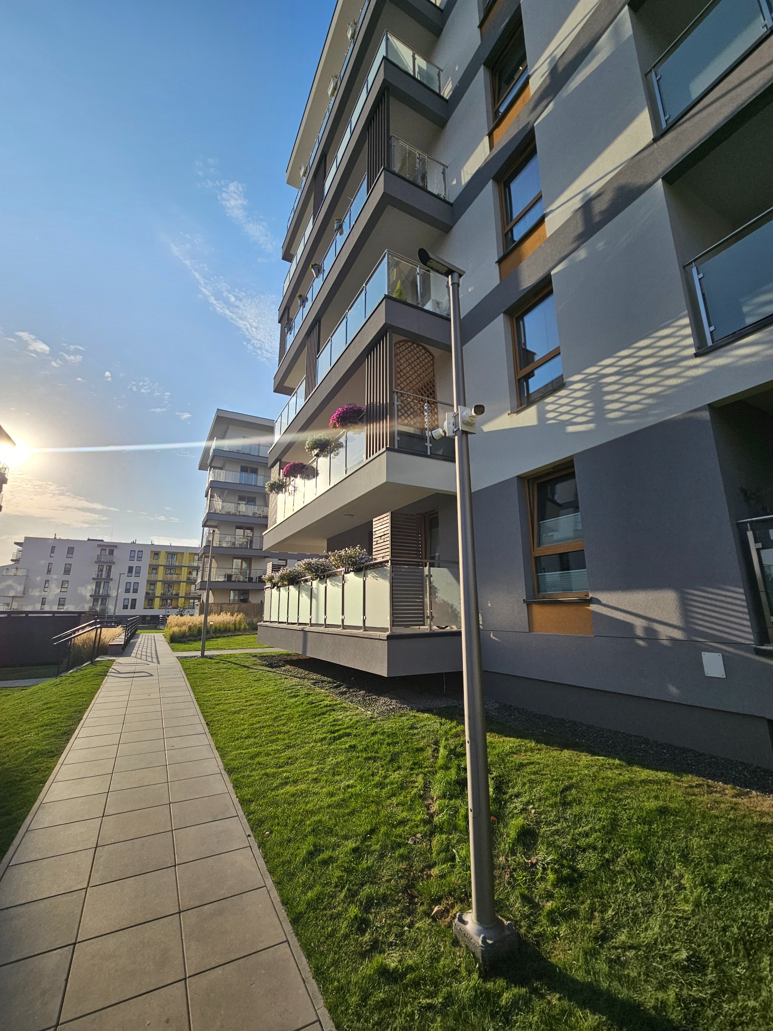 Kamera monitoringu zamontowana na słupie oświetleniowym przed nowoczesnym budynkiem mieszkalnym z balkonami i zielonym trawnikiem.