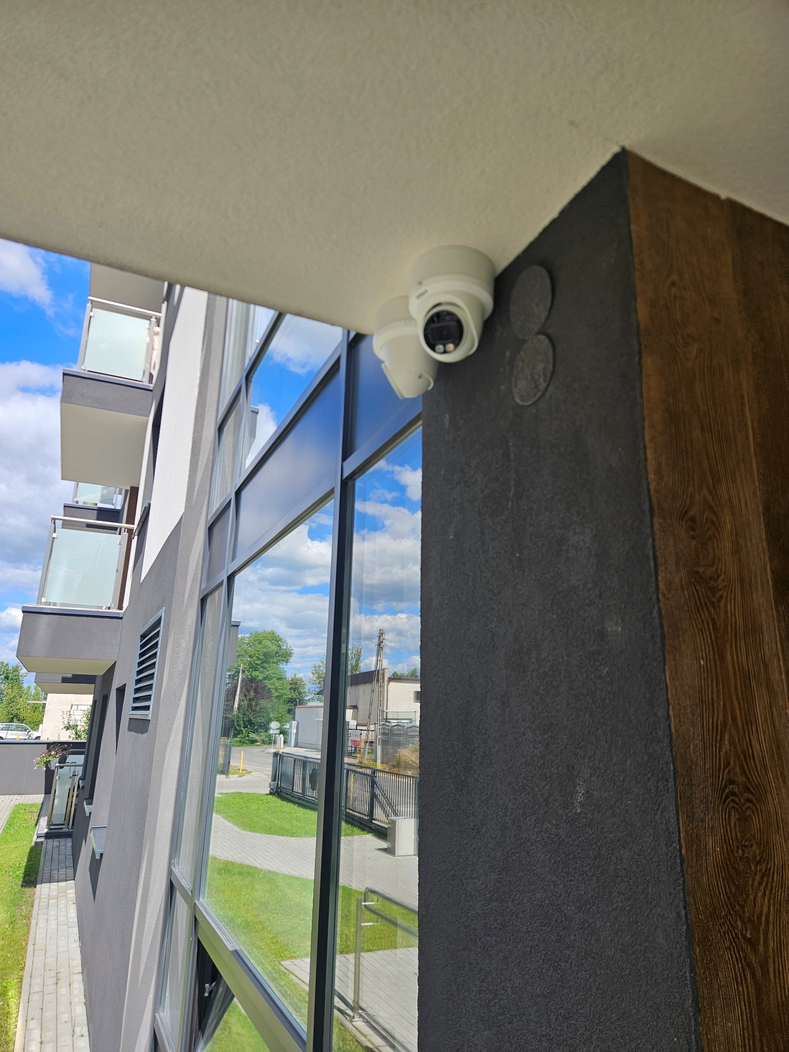Kamera kopułkowa zamontowana pod sufitem na rogu budynku z widokiem na otoczenie, widoczne okna z odbiciem nieba i fragment balkonu