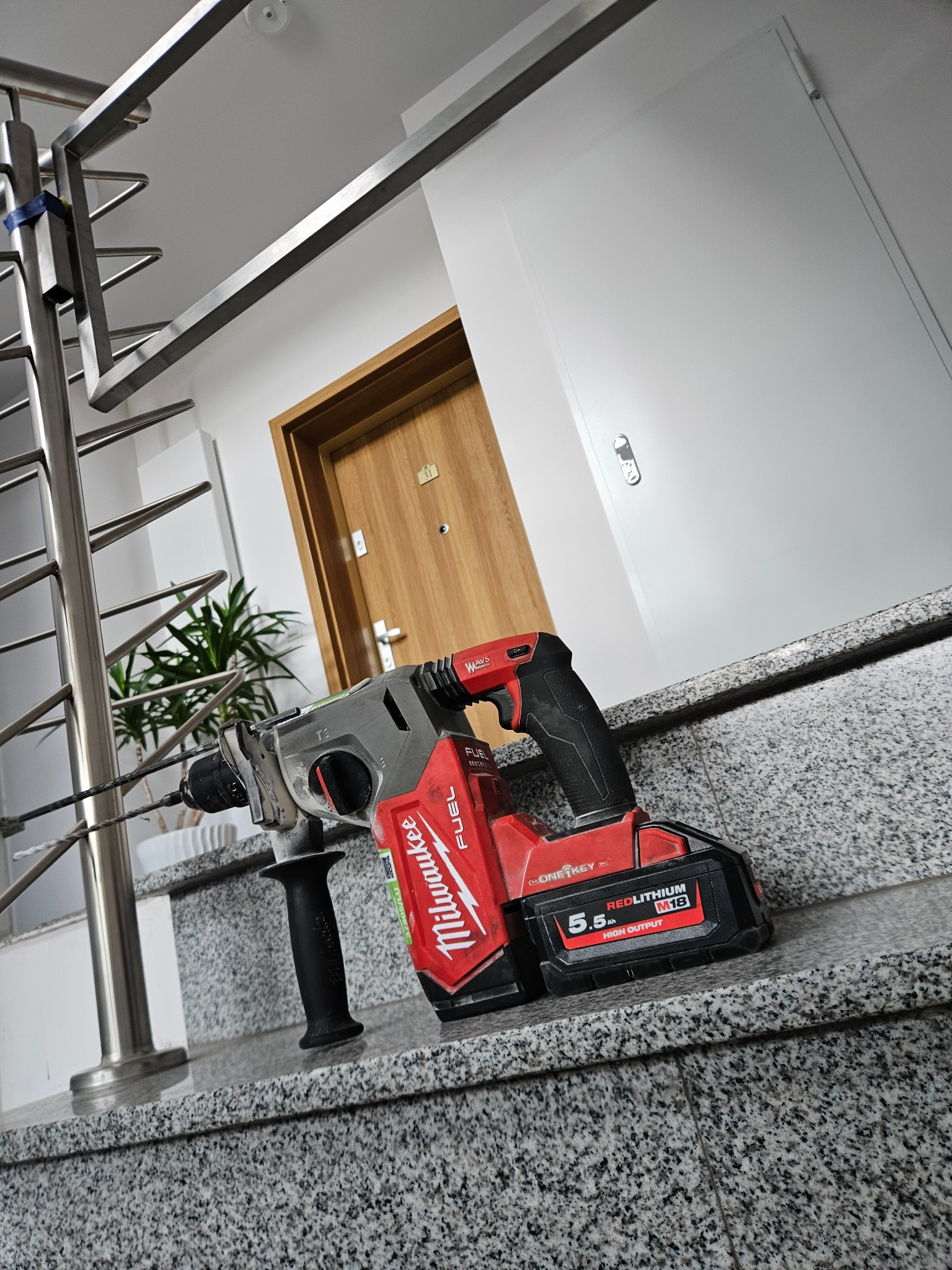 Akumulatorowa wiertarko-wkrętarka udarowa Milwaukee z zamocowanym wiertłem i dodatkową rękojeścią, leżąca na marmurowym parapecie w klatce schodowej, widok na stalową balustradę, drzwi wejściowe...