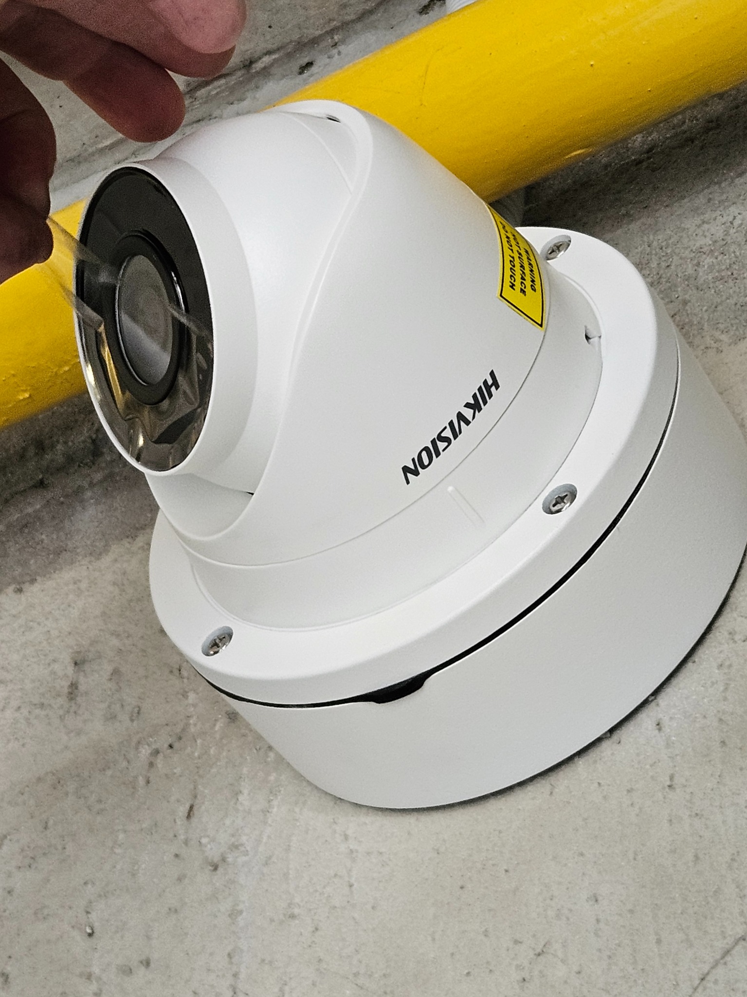 Biała kamera kopułkowa Hikvision, zdejmująca folię ochronną z obiektywu, zamontowana pod żółtą rurą na szarym, betonowym suficie.