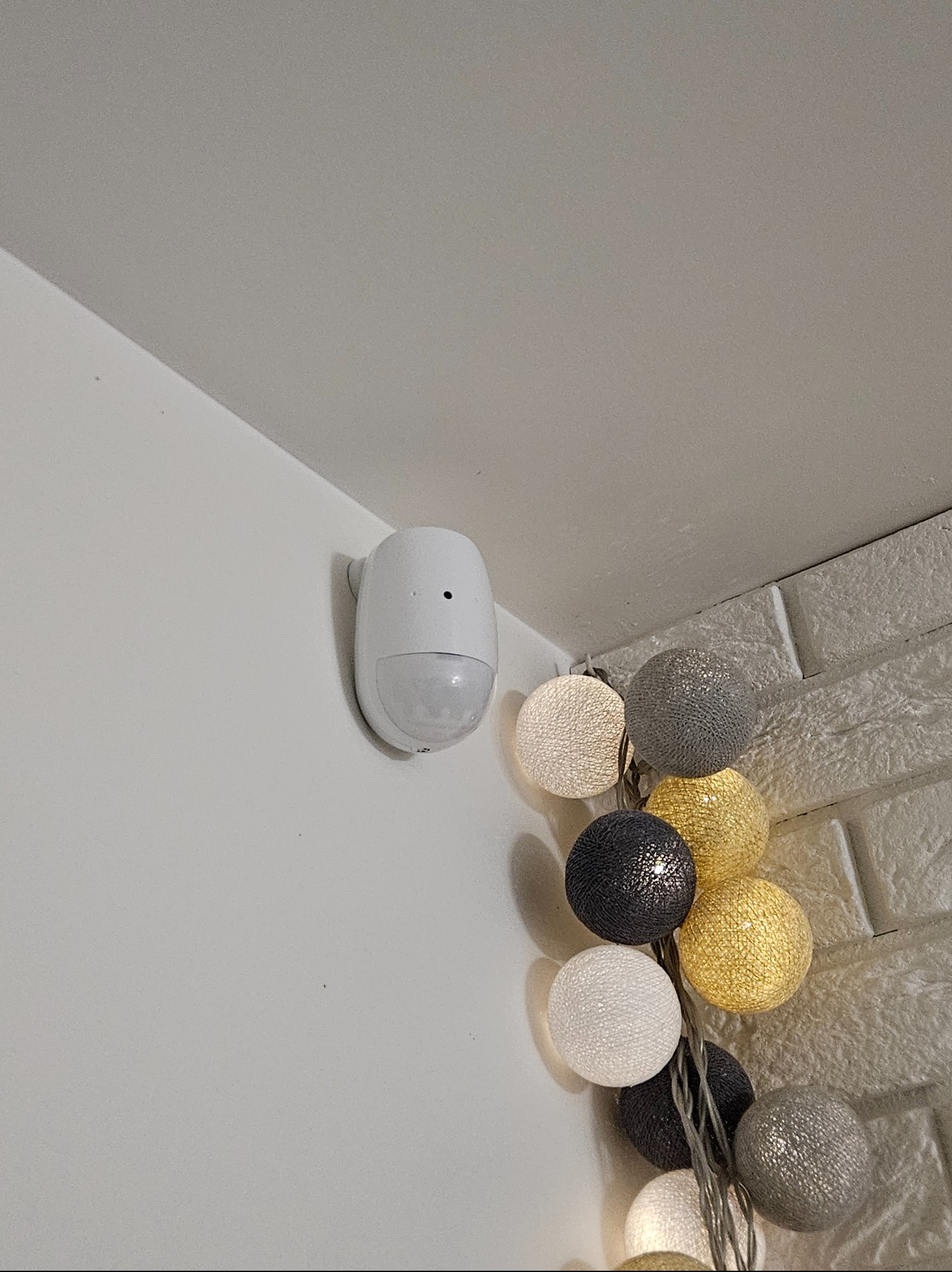 Biały czujnik ruchu zamontowany na ścianie, obok dekoracyjnych, świecących kul. Widok z dołu, oświetlenie ciepłe, wnętrze domu.