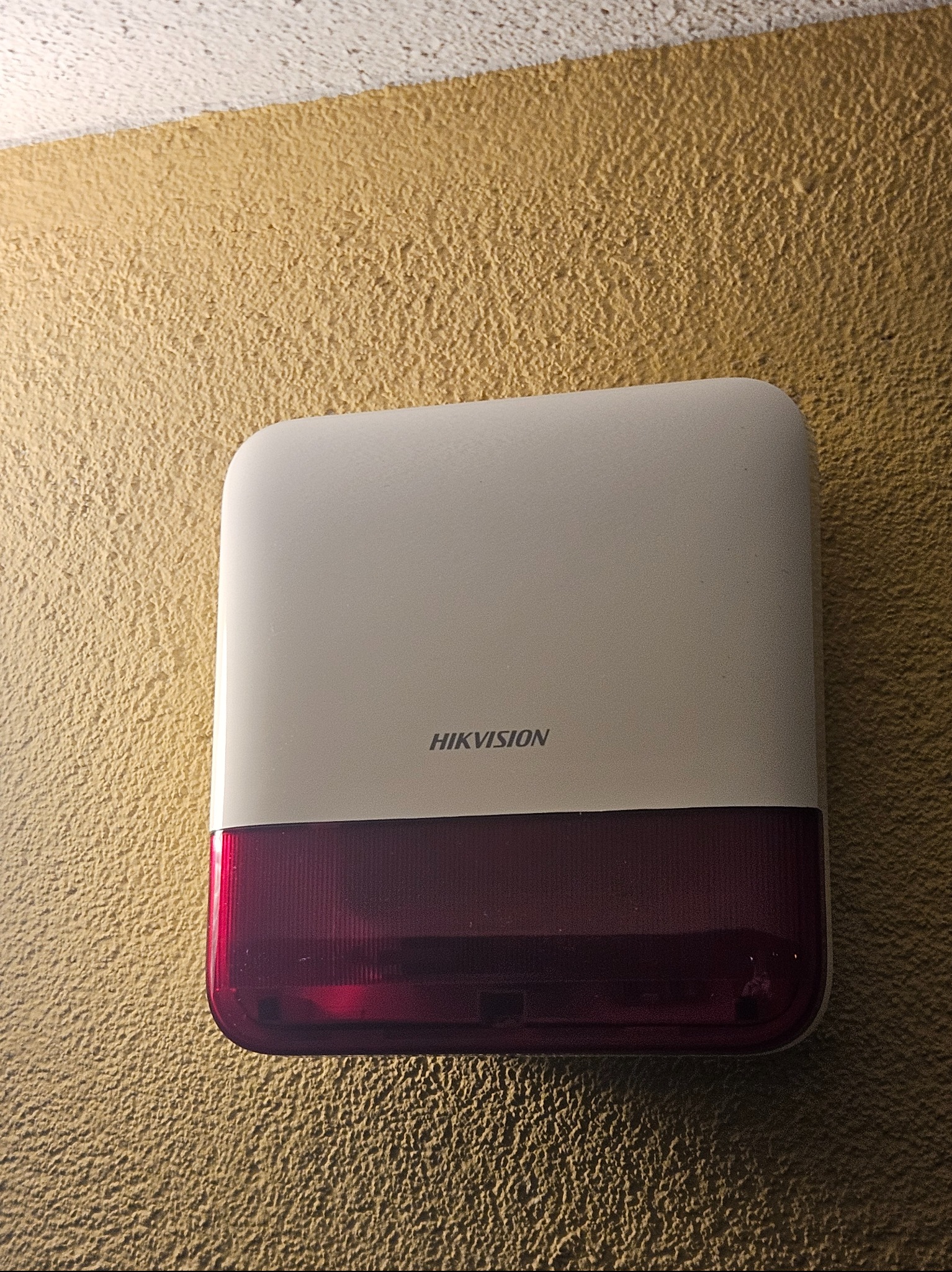 Zamontowana zewnętrzna syrena alarmowa Hikvision z czerwonym światłem na teksturowanej, beżowej ścianie. Widoczne logo producenta. Oświetlenie podkreśla fakturę ściany.