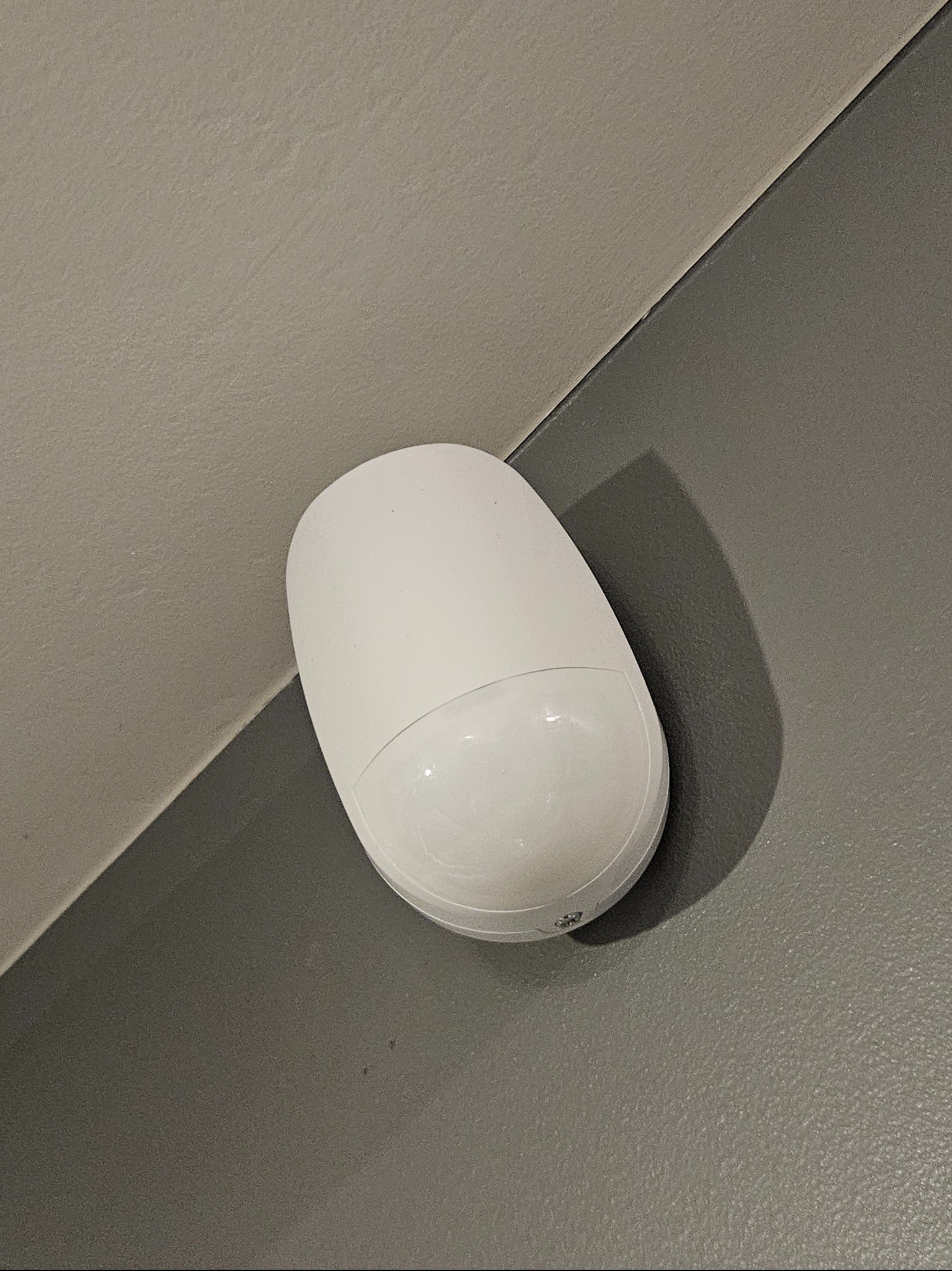 Zamontowany, biały czujnik ruchu alarmowego na styku dwóch ścian o różnych odcieniach szarości. Widoczny z bliska, z delikatnym cieniem.