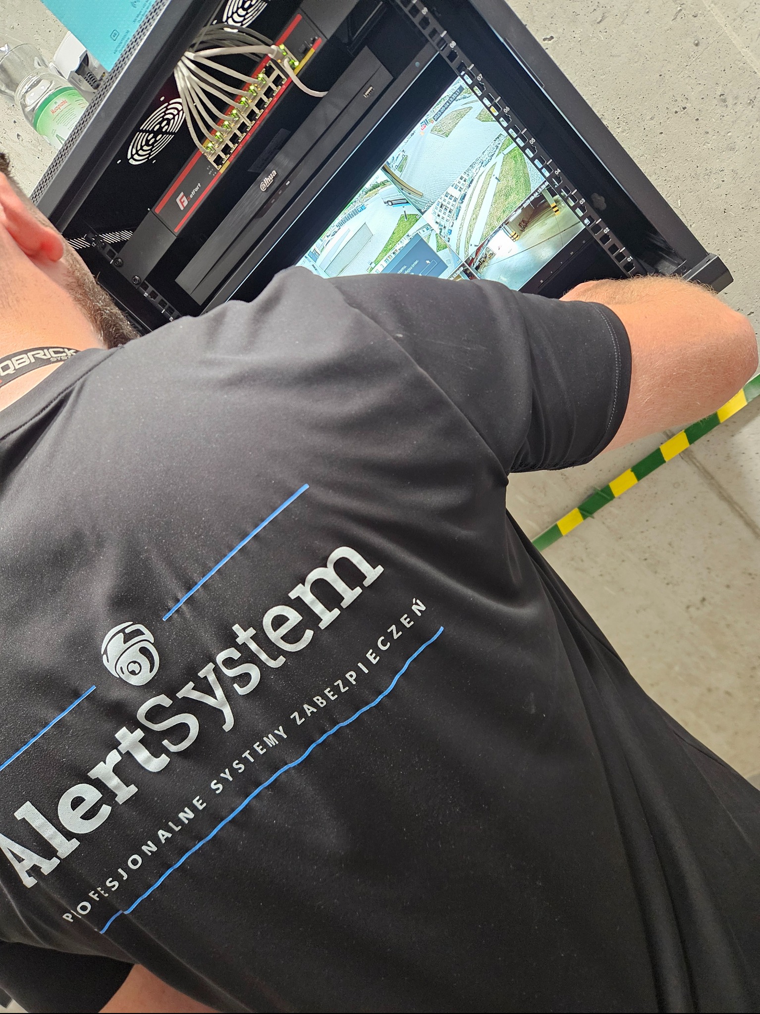 Instalator w czarnej koszulce z logo firmy montuje system kamer. Widoczny switch, rejestrator i podgląd z kamer na ekranie w szafie rack.