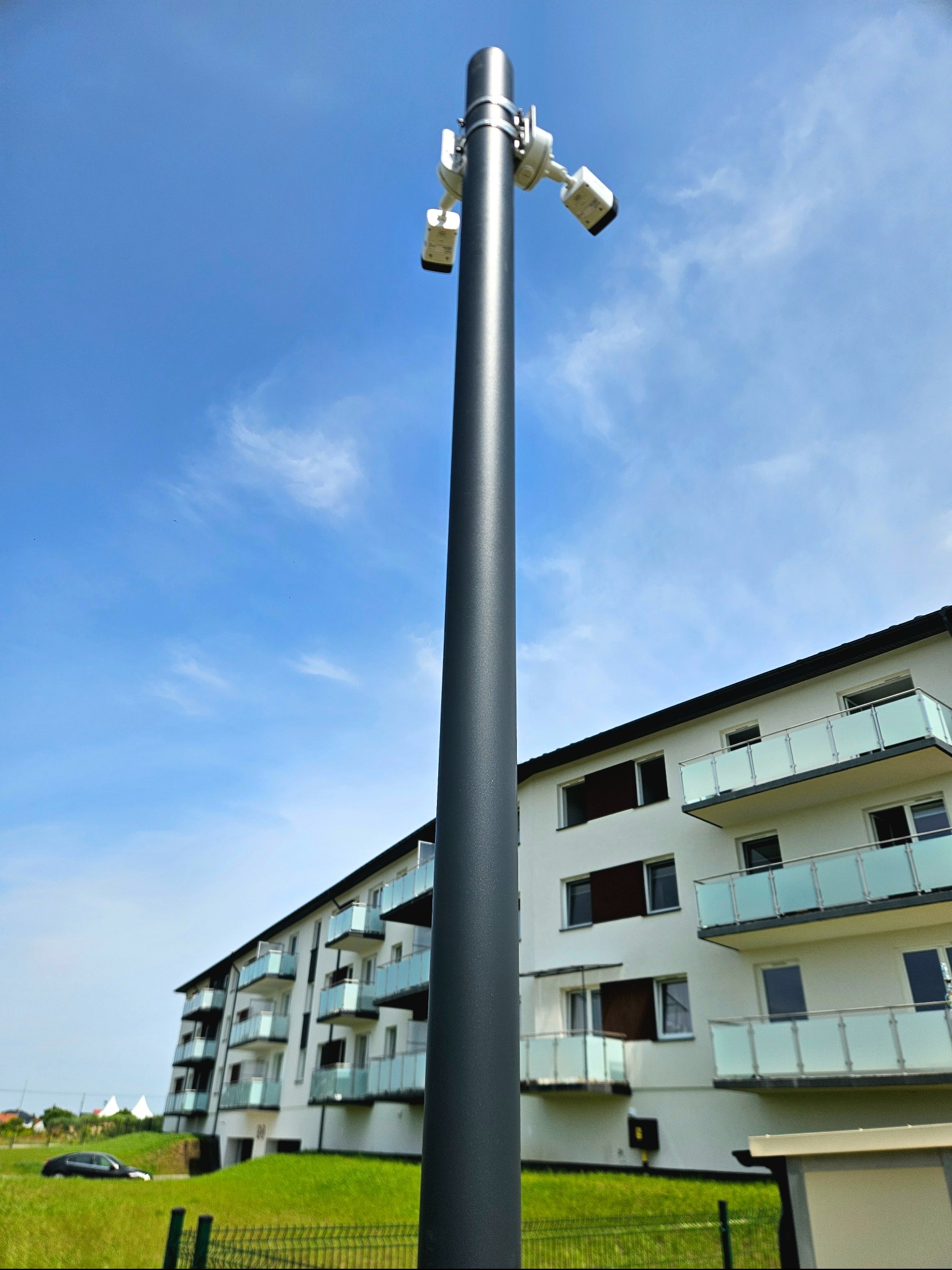 Słup z trzema kamerami monitoringu na tle błękitnego nieba i nowoczesnego apartamentowca z balkonami. Ujęcie z dołu, podkreślające wysokość słupa.