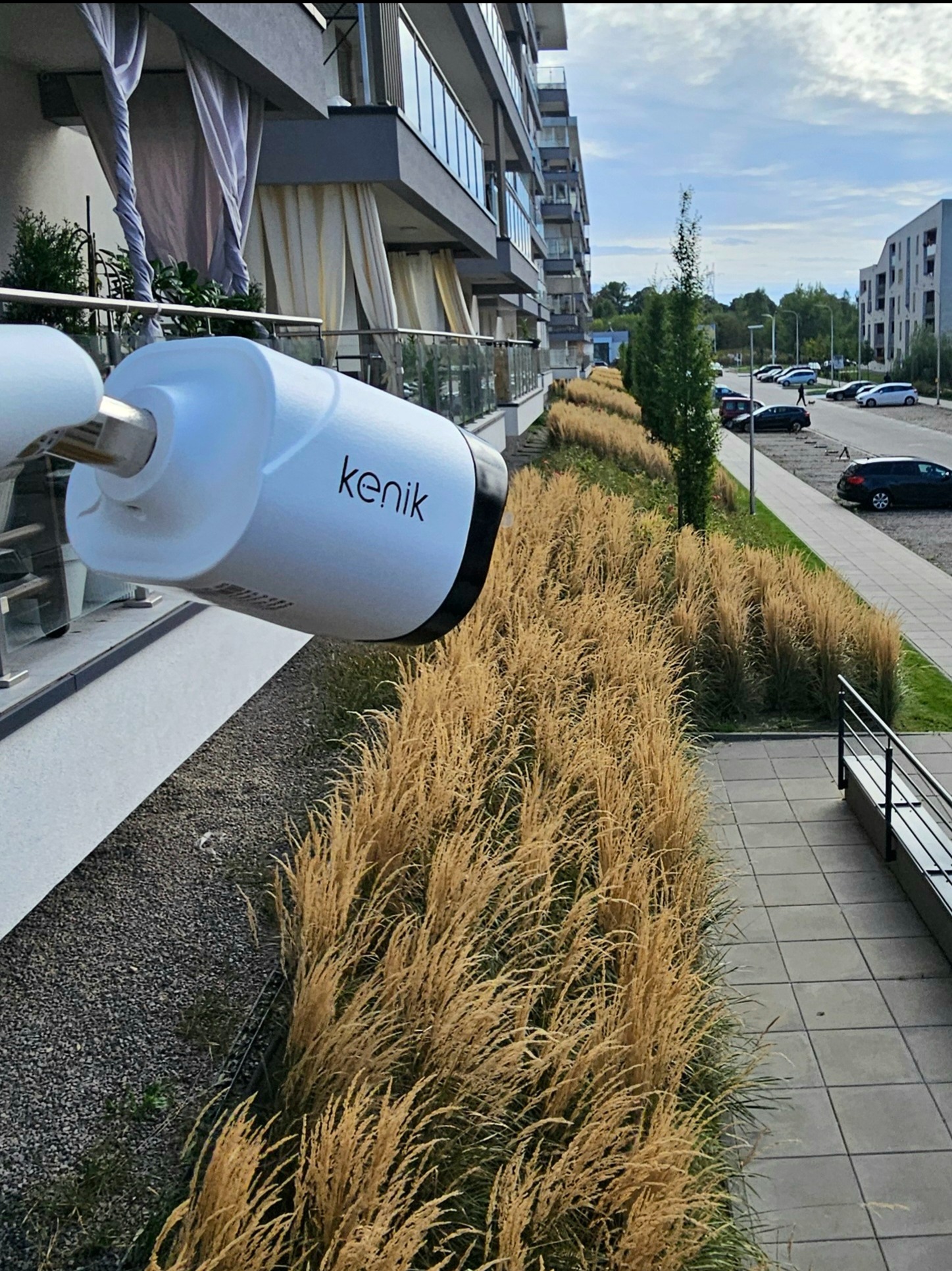 Kamera monitoringu marki Kenik zamontowana na balkonie nowoczesnego apartamentowca, z widokiem na parking i ozdobne trawy. Nietypowy kadr z bliska.