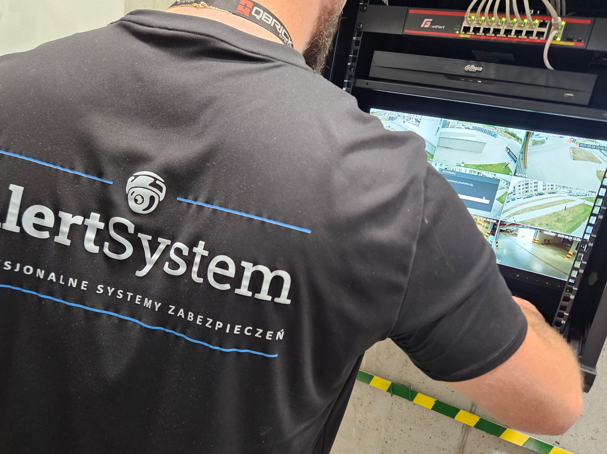 Pracownik w koszulce AlertSystem konfiguruje system monitoringu na monitorze z widokiem z kamer. Widoczny switch sieciowy i rejestrator Dahua.