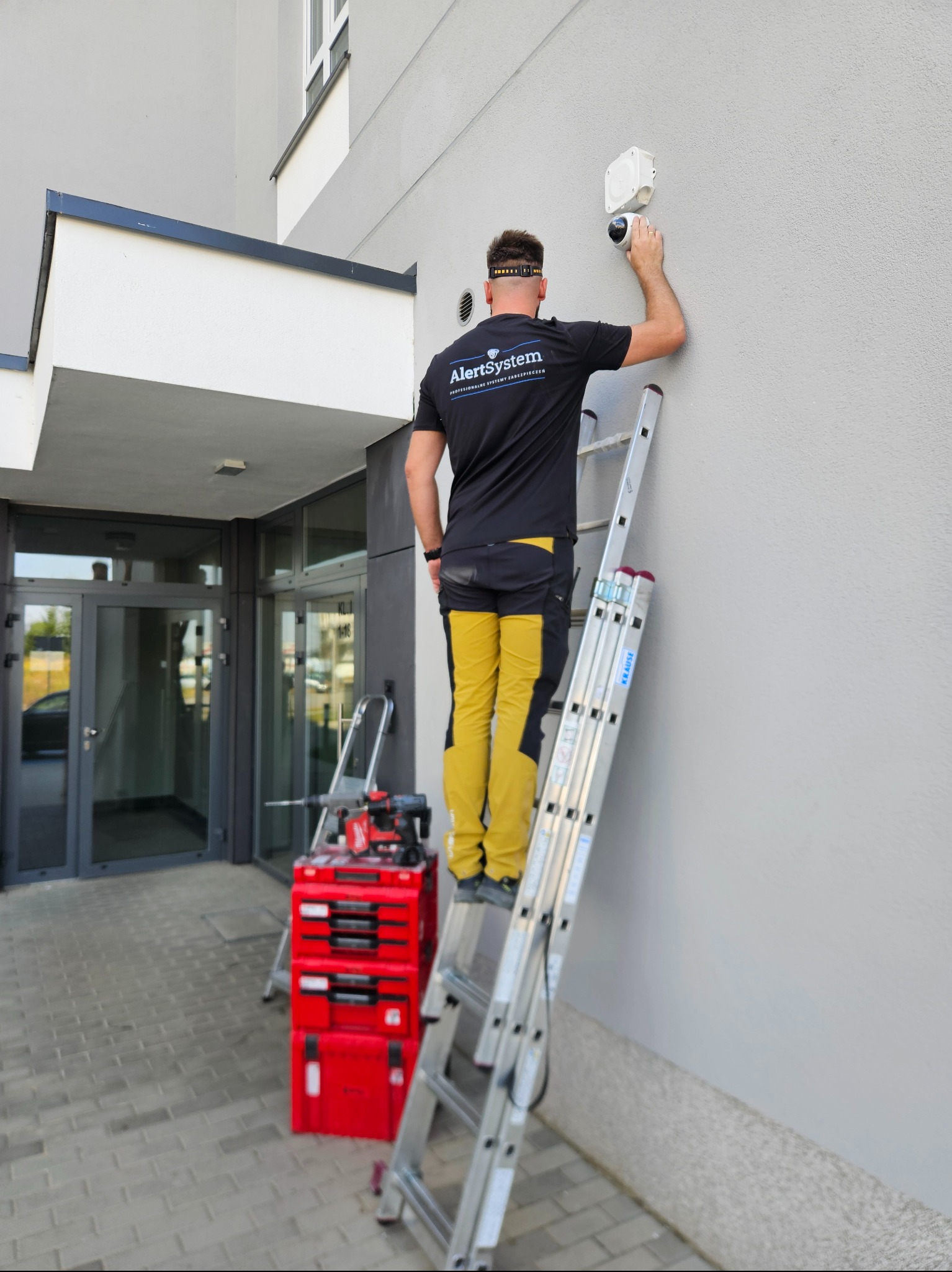 Instalator na drabinie montuje kamerę na elewacji budynku. Widoczna skrzynka z narzędziami i logo firmy na koszulce montera.