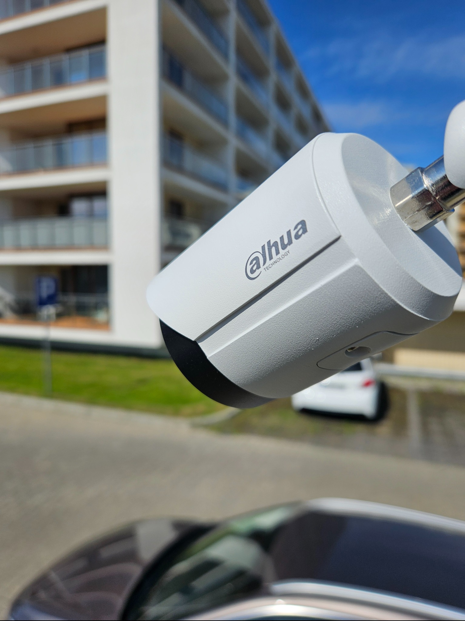 Kamera monitoringu Dahua na tle nowoczesnego budynku mieszkalnego i błękitnego nieba. Widoczny fragment samochodu w dolnej części kadru.