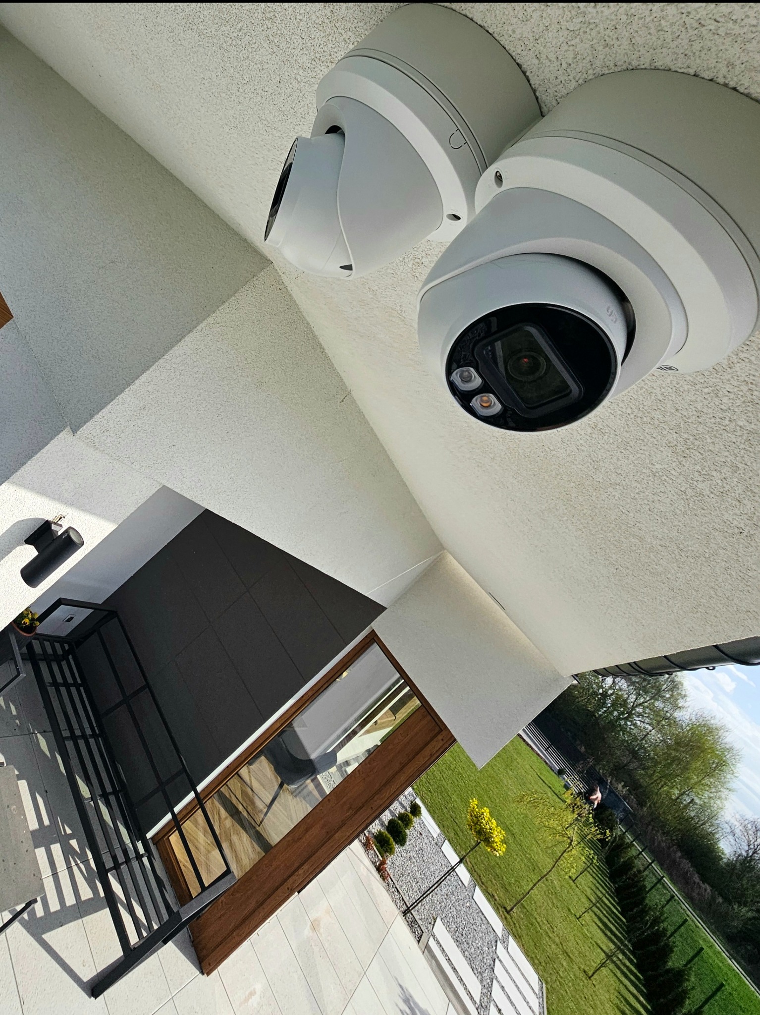 Dwie kamery monitoringu kopułkowe zamontowane na suficie budynku, widok z dołu, z fragmentem elewacji i ogrodu w tle. Kąt ujęcia podkreśla nowoczesny design.
