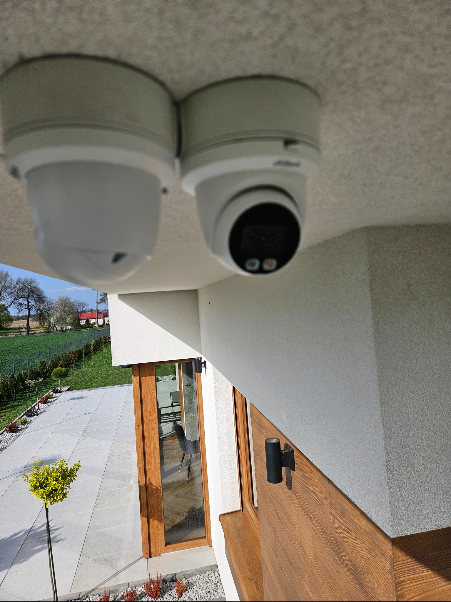 Dwie kamery kopułkowe zamontowane pod sufitem na zewnątrz budynku, widok na ogród i taras. Jedna kamera marki Dahua. Oświetlenie zewnętrzne na elewacji.