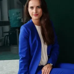 Portret kobiety w kobaltowym garniturze, siedzącej na podłodze, z delikatnym uśmiechem i zegarkiem na ręku, na tle zielonej ściany i biurowego wnętrza.