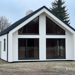 Solitar Fabryka Domów - Biały, parterowy dom z czarnym dachem i dużymi oknami, widoczny z zewnątrz na tle drzew i pochmurnego nieba. Nowoczesna architektura z minimalistycznym designem.